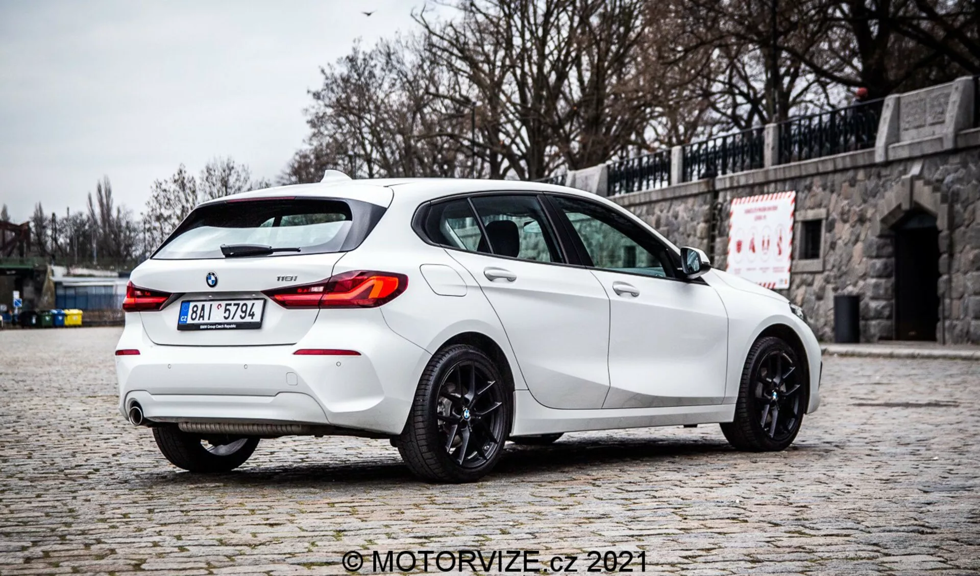 Visokokakovostna fotografija belega BMW 1 Series 2019 128ti hatchbacka, posneta iz zadnjega tričetrtinskega kota, poudarjajoč desno stran. Avto je parkiran na tlakovani površini s kamnitim zidom v ozadju, poudarjajoč njegov športni dizajn z zadnjim strešnim spojlerjem, dvojnim izpušnim sistemom, zadnjim difuzorjem, LED zadnjimi lučmi, zatemnjenimi okni in temno obarvanimi litimi platišči. Difuzna osvetlitev, odsotnost ostrih senc in jasne podrobnosti nakazujejo oblačno okolje, ki ponuja avtentično predstavitev barve in značilnosti vozila. Na sliki je viden vodni žig 'MotorVize.cz 2021'.