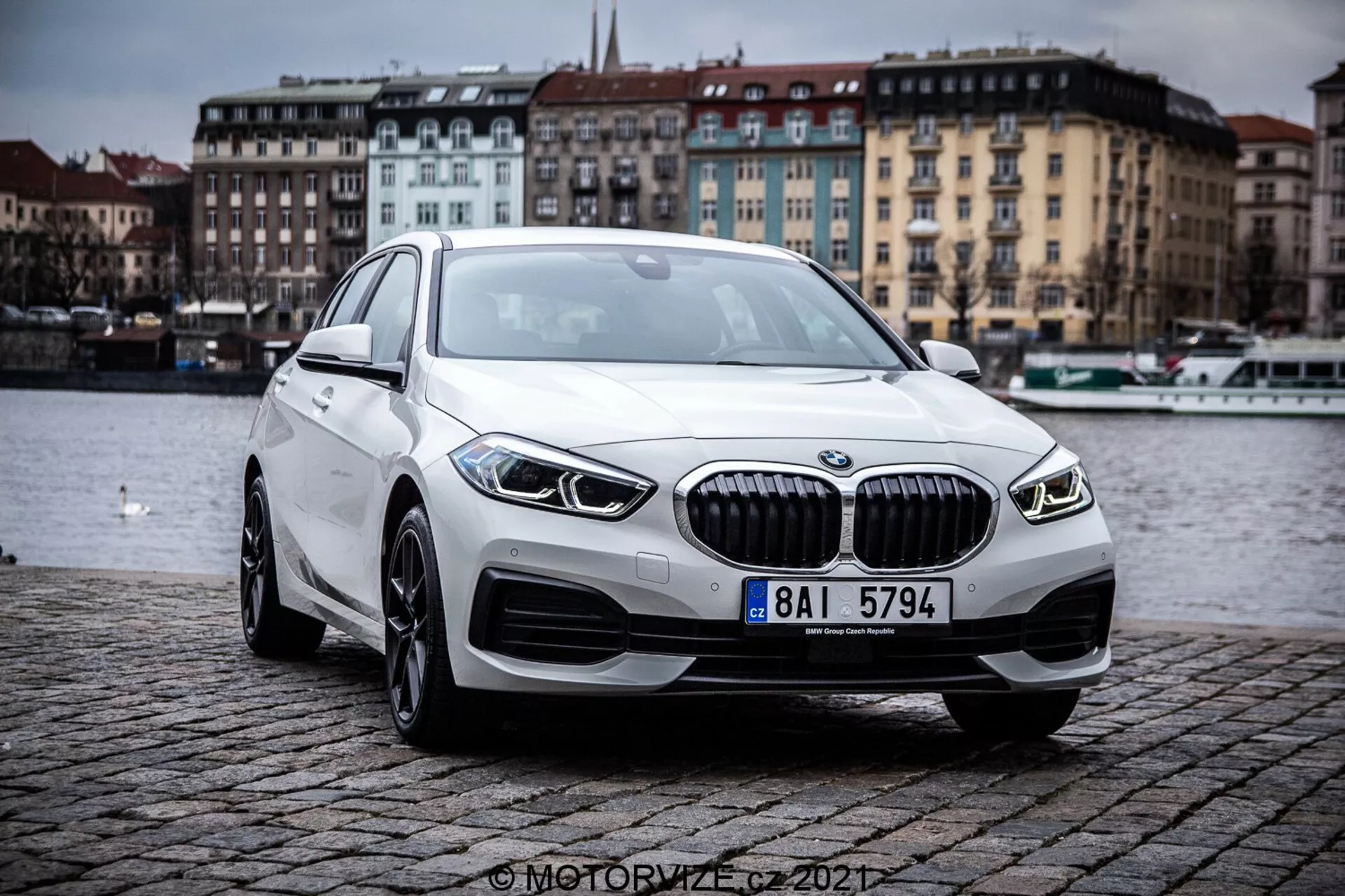 Slika prikazuje BMW 1er-Reihe 2019 (BMW 116i) iz treskrotnega sprednjega kota, ki poudarja obrise in oblikovalske elemente modernega luksuznega sedana. Avto se nahaja na prostem na tlakovani brežini ob vodnem telesu, pod oblačnim nebom. Pomembne značilnosti vključujejo mrežo z navpičnimi letvicami, elegantne LED žaromete, integrirane meglenke, aluminijasta platišča in aerodinamični profil.