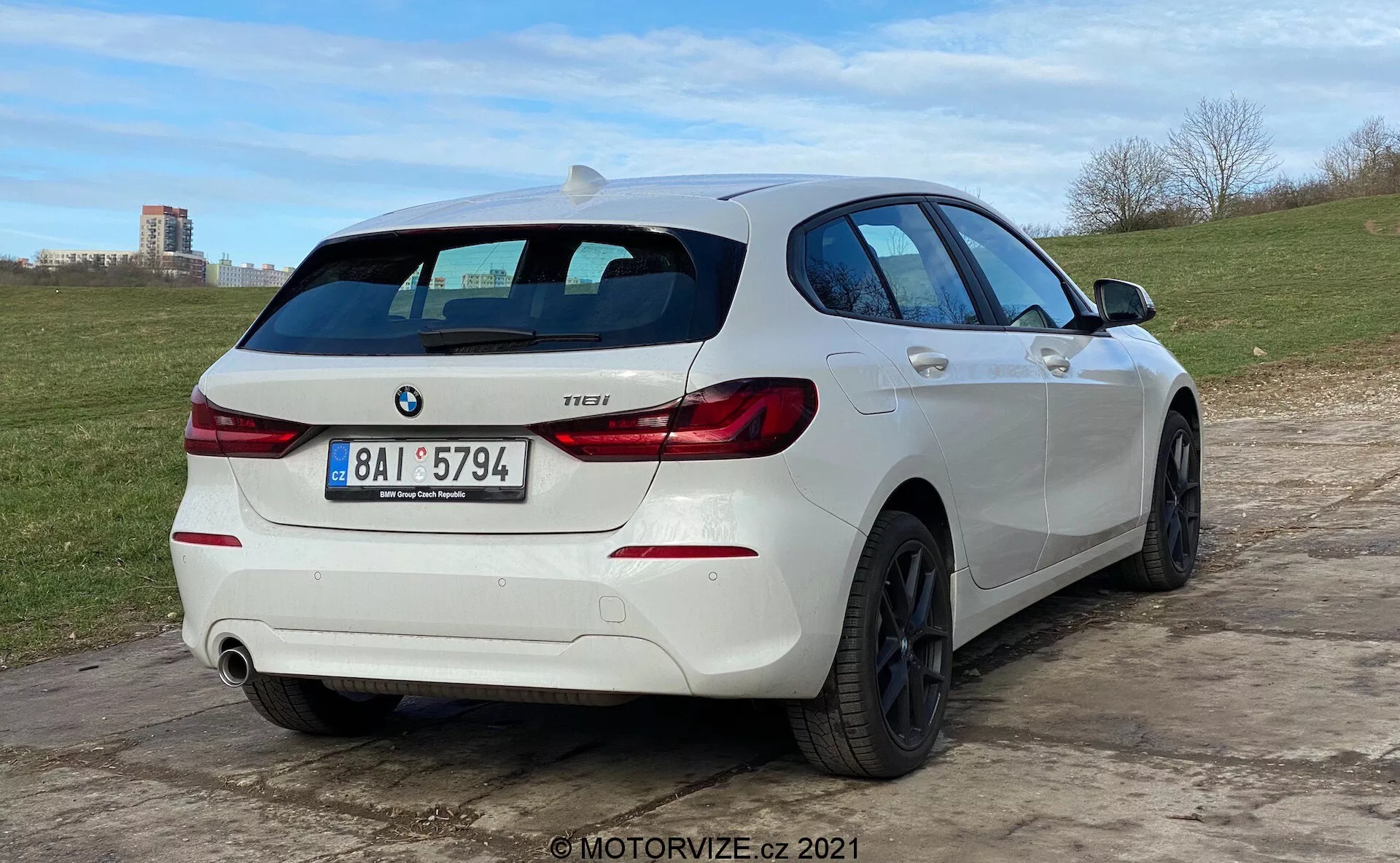 Pogled iz treh četrtin od zadaj na beli BMW 1 Series 118i hatchback, parkiran na neenakom tleh. Avto ima značilne zadnje luči v obliki črke 'L', značko BMW in značko modela z oznako '118i'. Ima dvojni izpušni sistem z eno vidno cevjo na desni strani zadnjega odbijača. Vozilo je opremljeno z večkrakimi črnimi aluminijastimi platišči in zadnjim brisalcem. Strešni spojler dodaja športni videz, stranski profil pa razkriva rahlo zatemnjena okna, gube na karoseriji ter ročaje in ogledala, ki ustrezajo barvni shemi avta. Nizek kot kamere poudarja držo avta in oblikovanje koles.