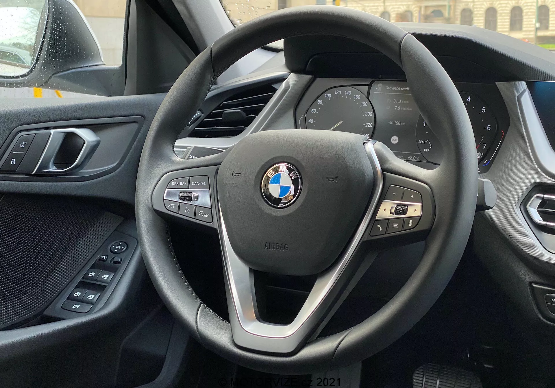 Pogled v notranjost BMW serije 1 2019 (BMW 116i) iz voznikovega sedeža s poudarkom na sodobnem trikraku BMW volanu z gumbi za nadzor multimedije, telefona in tempomata ter digitalni instrumentni plošči, ki prikazuje ključne podatke o vozilu, kot sta hitrost in vrtljaji. V ozadju je videti zgornji rob sredinske konzole, levo pa je viden del voznikovih vrat z električnimi upravljalnimi elementi za udobje. Armaturna plošča in vratna panelna plošča prikazujeta kombinacijo mehkih materialov in kakovostne izdelave. BMW znak in iPERFORMANCE značko sta jasno vidna na volanu, kar nakazuje na učinkovitost ali hibridno tehnologijo.