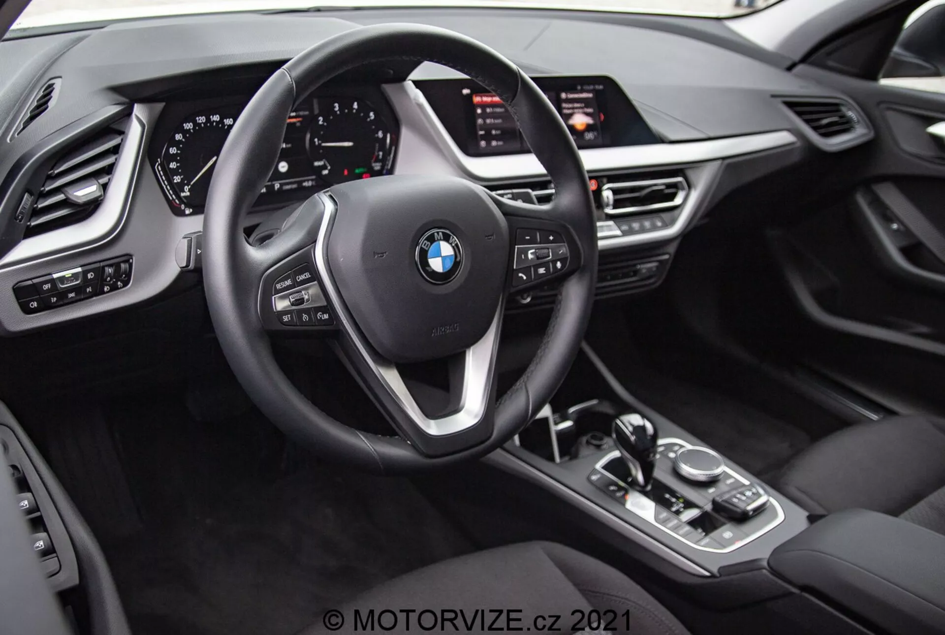 Pogled na notranjost BMW 1er-reihe 2019 (BMW 116i) iz perspektive voznika. Slika poudarja BMW-emblem na trikrakam volanu z večfunkcijskimi gumbi in obvolanskimi ročicami. Za volanom je popolnoma digitalni instrumentni grozd z osvetljenimi številčnicami in različnimi informativnimi prikazi. Sredinska konzola vsebuje prestavno ročico avtomatskega menjalnika, upravljalni gumb iDrive, gumbe za načine vožnje in velik infotaiment zaslon, nekoliko nagnjen proti vozniku. Notranjost vključuje teksture, ki posnemajo usnje, kovinske poudarke in dodatne kontrole za žaromete, okna in nastavitve ogledal s funkcijami spomina za voznikov sedež.