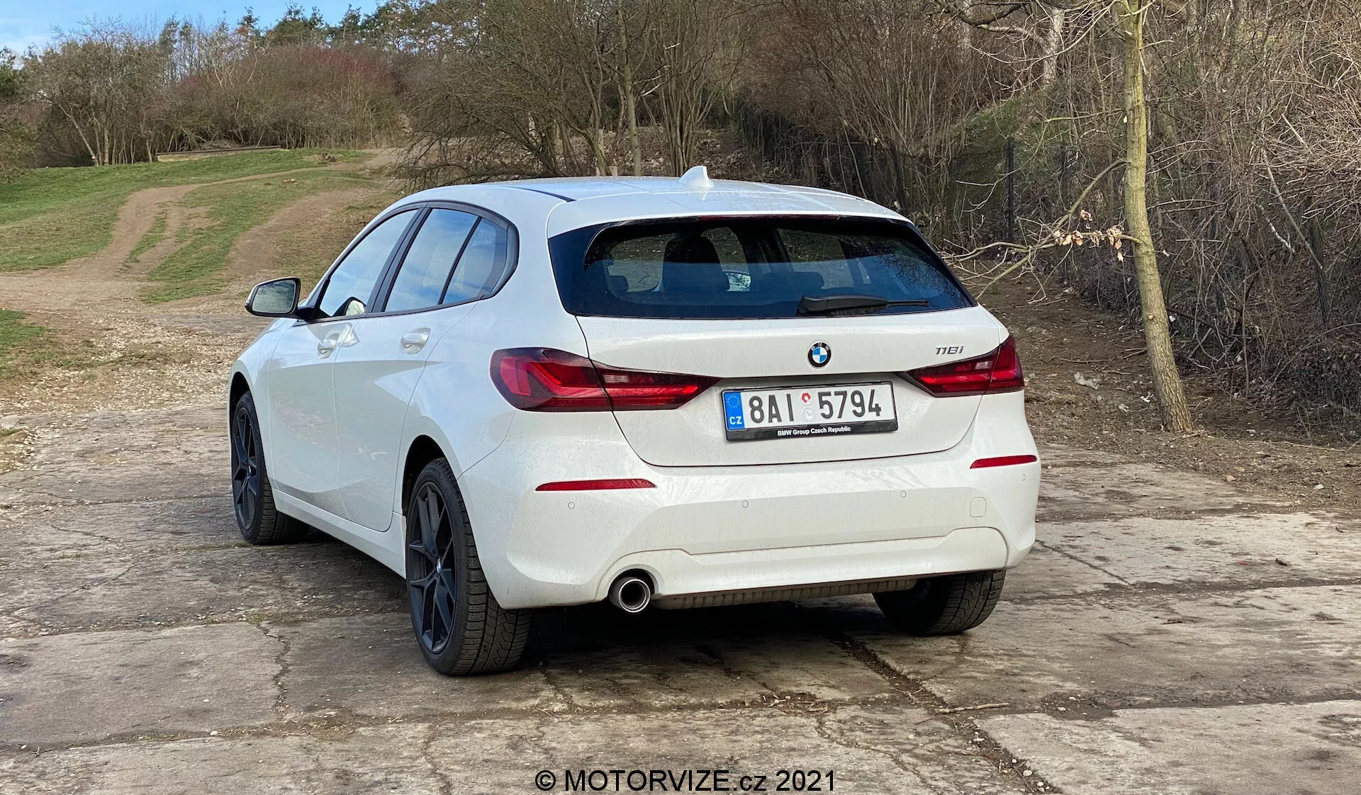 Slika prikazuje belega BMW 1er-reihe 2019 (BMW 116i) hatchback iz kota zadnje tretjine vozila. Zadnji del vozila, vključno s horizontalno nameščenimi zadnjimi lučmi, dvojnim izpušnim sistemom in parkirnimi senzorji, vgrajenimi v zadnji odbijač, je jasno viden. Na zadnjih vratih je videten BMW logotip in oznaka modela. Avto je postavljen na gramozni površini z naravnim, podeželskim ozadjem. Slika nakazuje avtomobilske teme, kot so oblikovanje vozil, sodobni modeli avtomobilov, vsebina specifična za BMW, izpušni sistemi in tehnologija za pomoč pri parkiranju. Prisotnost vodnega žiga MOTORVIZE.cz 2021 kaže na izvor v avtomobilskem pregledu ali publikaciji.