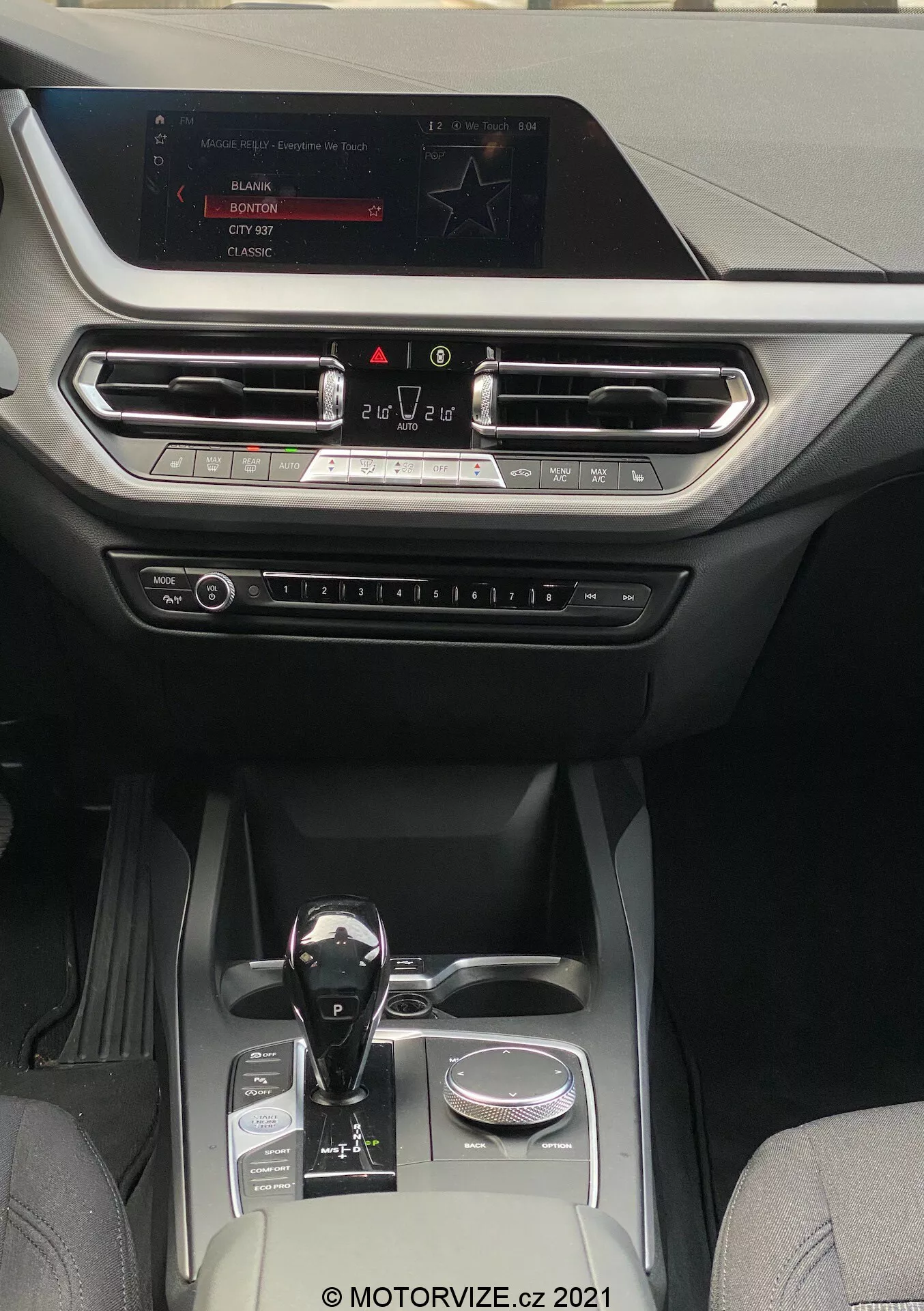 Pogled v notranjost BMW 1er-reihe 2019 (BMW-116i), ki prikazuje moderno in luksuzno kabino, osredotočeno na osrednjo konzolo in področje armaturne plošče. Perspektiva je iz območja zadnjih sedežev, obrnjena naprej proti zaslonu infotainment, nameščenemu na vrhu armaturne plošče. Zaslon prikazuje možnosti za radio, medije in nastavitve avtomobila. Pod zaslonom so horizontalno poravnani zračniki s kovinskimi poudarki in dekorativnim trakom, ki sega čez celotno širino armaturne plošče. Pod zračniki so vidni fizični gumbi in gumbi za nadzor klimatske naprave in druge nastavitve. Samodejna prestavna ročica ima sijoč črni zaključek s kovinskimi poudarki, nameščena poleg vrtljivega gumba. Osrednja konzola vključuje dva držala za pijačo, majhen shranjevalni prostor in srebrni dekorativni element, kar kaže na dobro opremljeno kabino, ki združuje usnje in visokokakovostne sintetične tkanine, primerno za avtomobile srednjega do višjega razreda, ki se osredotočajo na tehnologijo in udobje.
