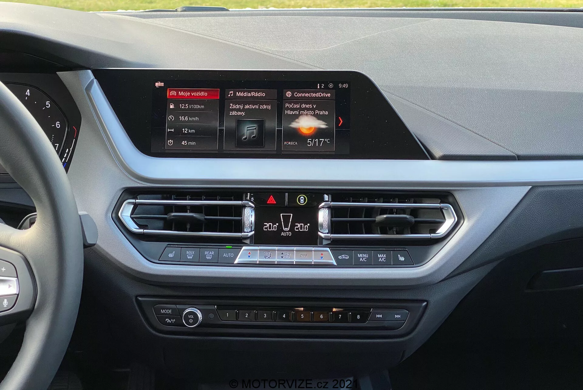 Notranji pogled na BMW serije 1 2019 (BMW-116i) z voznikove perspektive, prikazuje sredinsko konzolo armaturne plošče. V središču je pravokotni in osvetljen zaslon na dotik infotainment sistema, ki prikazuje navigacijo, medije in podrobnosti o klimi. Pod zaslonom so prezračevalne odprtine s kovinsko obrobo, zasilni kontrolniki in kroženje zraka, sledi panel za nadzor klime s temperaturnimi gumbi, hitrostjo ventilatorja in gumbi za ogrevane sedeže. Materiali imajo mehke površine in kromirane poudarke, kar kaže na osredotočenost na udobje in udobje voznika ter sovoznika.