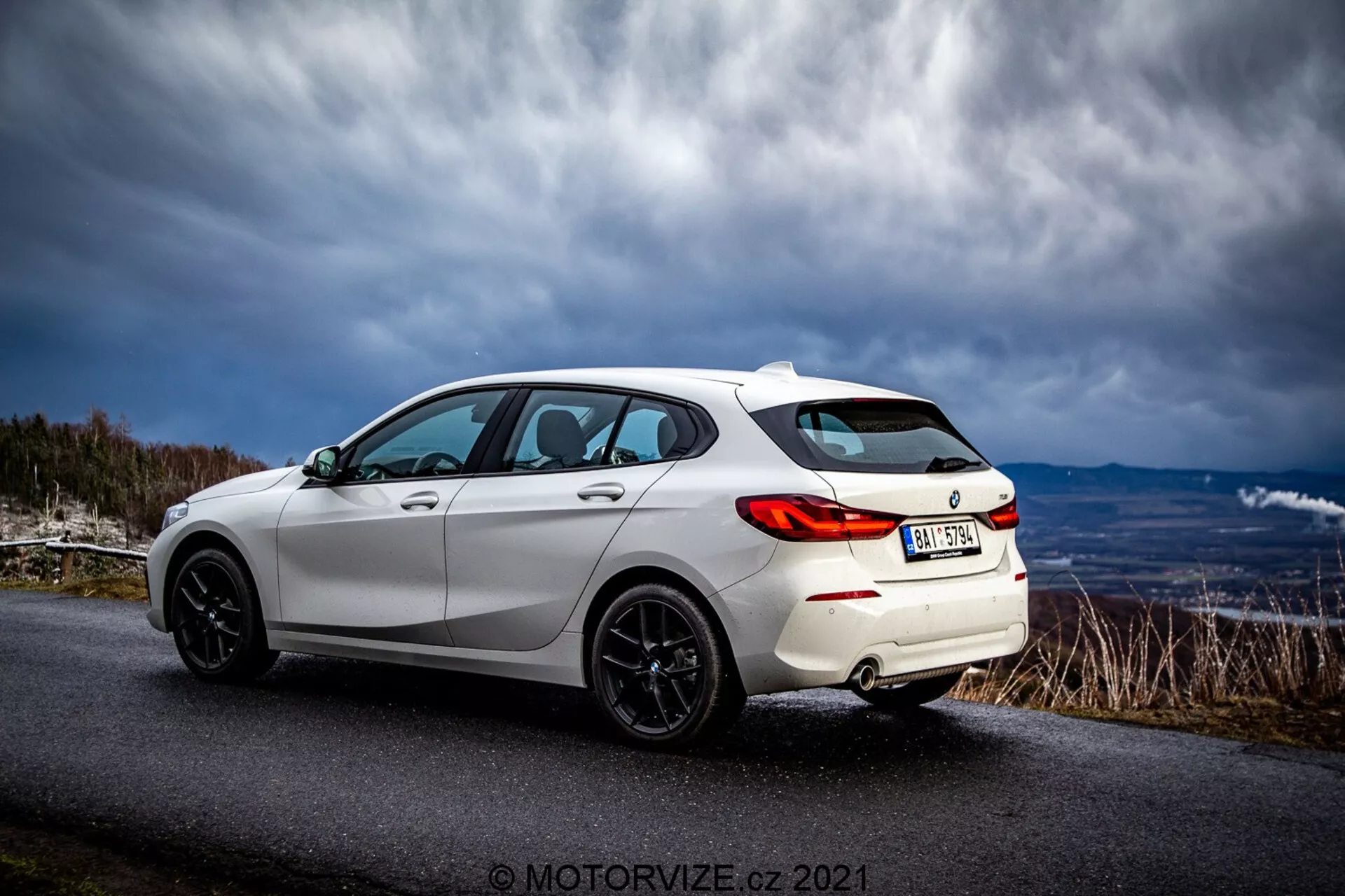 Bel, sodoben hatchback BMW 116i 2019, ujet z vidika treh četrtin od zadaj. Zunanje športne lastnosti vključujejo LED zadnje luči, črna večnapravčna lahka platišča, zadnji spojler, tonirana okna in elegantne linije karoserije, ki poudarjajo aerodinamični dizajn. Vozilo je parkirano na asfaltirani cesti v podeželskem ali gorskem območju pod dramatičnim nebom.