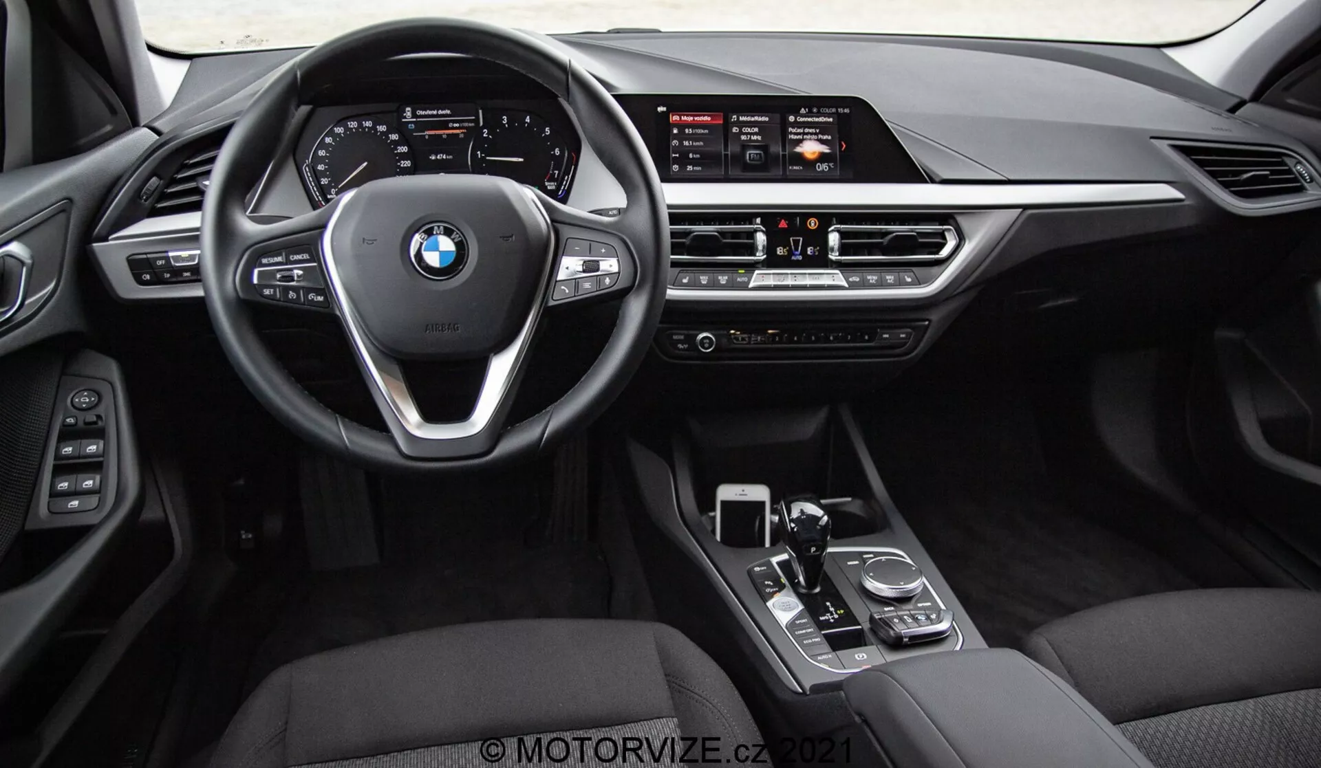 Pogled na notranjost BMW serije 1 2019 (BMW 116i), posnet iz zadnjih sedežev, prikazuje celotno sprednjo armaturno ploščo, volan z logotipom BMW in kontrolnimi gumbi ter sredinsko konzolo. Vidni elementi vključujejo širok infozabavni zaslon, ročne klimatske kontrole, zračne odprtine, prestavno ročico, vrtljivi gumb in sledilno ploščico. Notranjost je obdelana v črnem usnju ali umetnem usnju z kovinskimi in sijočimi zaključnimi elementi, poudarjajoč luksuz in napredno voznikovo vmesniško tehnologijo.
