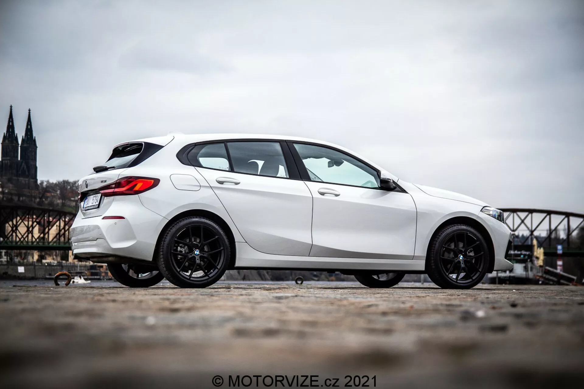Bel hatchback BMW serije 1 (BMW 116i) iz leta 2019, fotografiran z bočnega profila, ki prikazuje stran vozila od prednjega kolesa do zadnjega dela. Avto je postavljen na levi strani slike z zamegljenim zgodovinskim mestnim ozadjem, ki vključuje katedralo in most. Slika poudarja športna temna platišča avtomobila in njegovo dominantno držo zaradi nižje perspektive tal in rahle nagnjenosti kamere navzgor. Fotografija je posneta na prostem podnevi, kar razkriva podrobnosti estetskega videza avtomobila v naravni dnevni svetlobi.