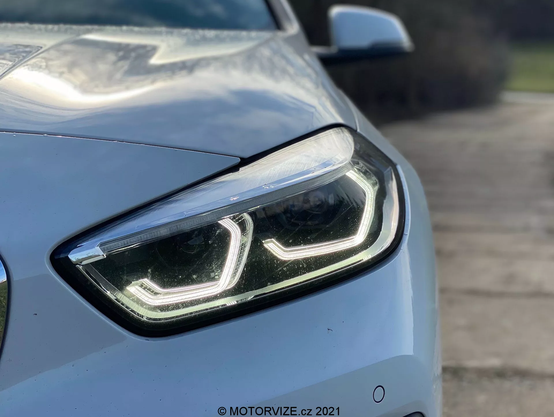 Približan posnetek serije BMW 1 2019 (BMW 116i) iz sprednjega kotnega kota, ki poudarja levo stran in se osredotoča na eleganten, moderen dizajn levega sprednjega žarometa z LED dnevnimi lučmi in prozornim pokrovom leče. Aerodinamične linije pokrova motorja in blatnika so delno vidne, kar prikazuje svetlo kovinsko barvo in sijoč zaključek avtomobila. Zamegljeno ozadje poudarja napredne tehnološke lastnosti žarometa, kar nakazuje LED ali HID svetilke, morda opremljene z adaptivno tehnologijo osvetlitve.