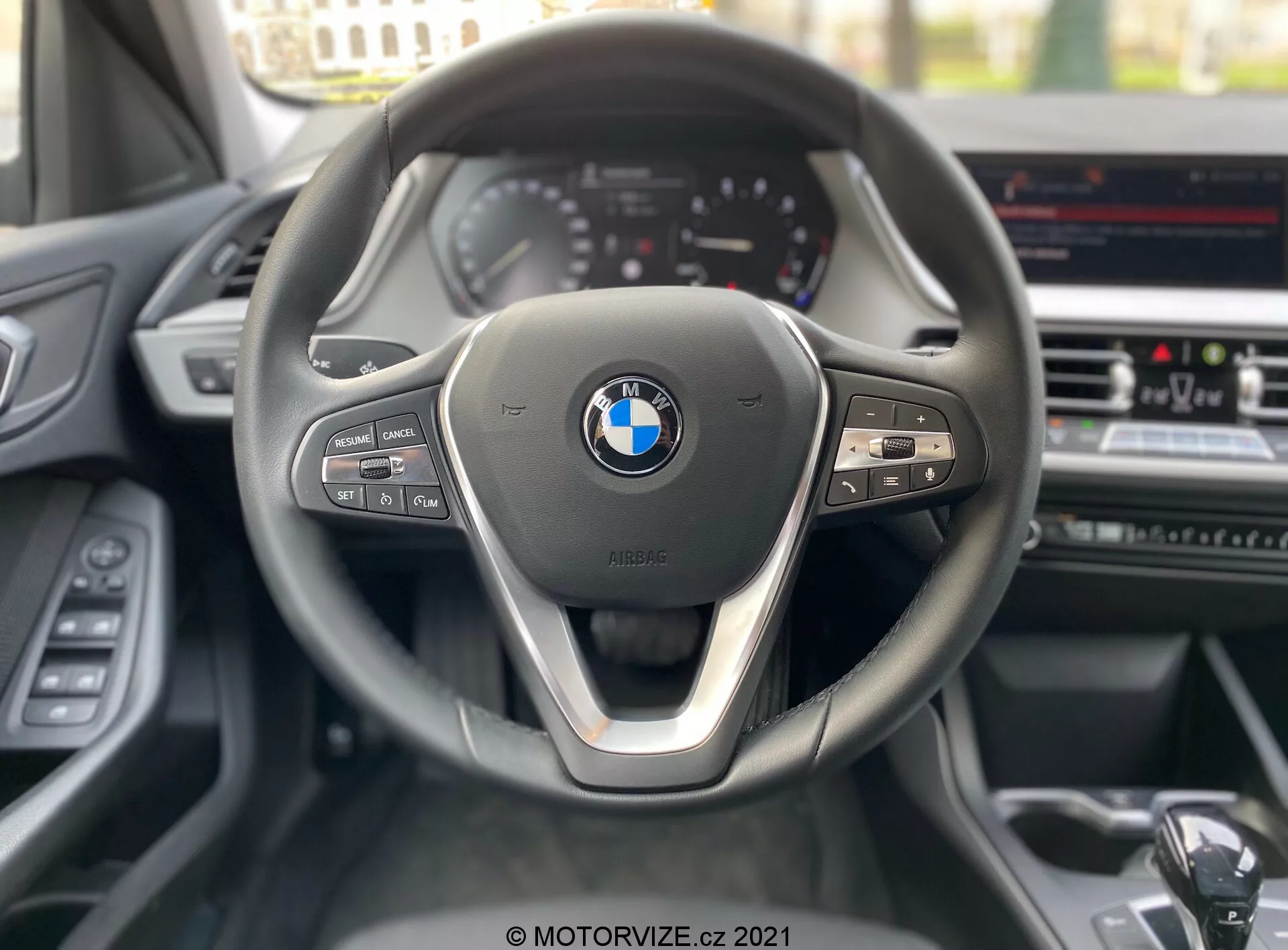 Perspektiva z voznikovega sedeža BMW serije 1 2019 (BMW-116i), osredotočena na sodoben trikraki volan s kovinskim poudarkom na spodnji kraki in vidnim BMW emblemom v središču. Na levi strani volana so kontrole za medije in komunikacijo, medtem ko so na desni strani gumbi za tempomat in vidna prestavna ročica. V ozadju je delno viden instrumentni sklop z analognimi merilniki in zaslon za informacije in zabavo na sredinski konzoli, kar nakazuje na napredne funkcije, kot so GPS navigacija in klimatska naprava.
