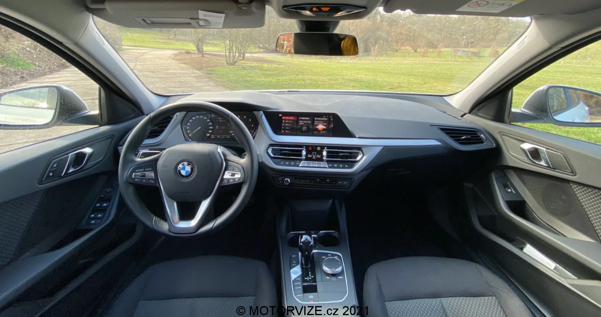 Pogled iz notranjosti s zadnjega sedeža BMW serije 1 2019 (BMW-116i), ki prikazuje sodobno kabino s trifunkcijskim volanom z logotipom BMW, samostoječim zaslonom za informiranje, dvoobmočno klimatsko napravo in avtomatskim menjalnikom. Uporaba usnja, mehkega plastika in kovinskih poudarkov poudarja visoko raven opreme.