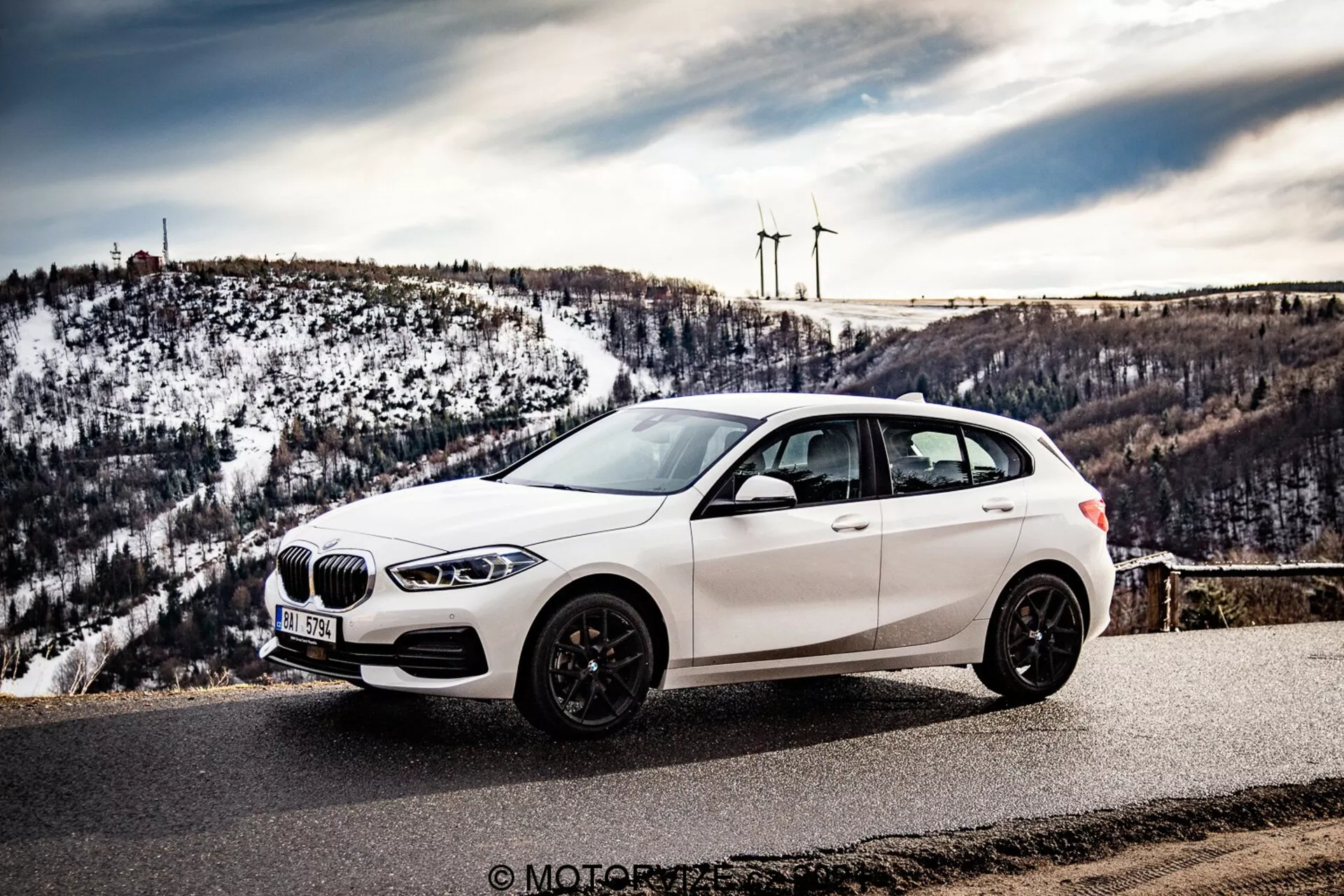 Slika prikazuje bel hatchback BMW 116i 2019, parkiran na slikovitem razglednem mestu ali gorski cesti, fotografiran iz bočno-sprednjega kota, ki prikazuje sprednjo masko, LED žaromete, prednje kolo, bočni profil in pogled na zadnji del. Perspektiva je rahlo zvišana, kar poudarja streho avtomobila, ki je pomaknjena v levo, v ozadju pa so zasneženi hribi in oddaljene vetrne turbine. Vozilo ima ledvično masko, nizkoprofilne pnevmatike na večkrakih aluminijastih platiščih in temna platišča, stranska ogledala z vgrajenimi smerokazi in zatemnjena okna. Okolica nakazuje jasno vreme, kar poudarja promocijsko in vizualno privlačnost fotografije, ki je bila morebiti posneta med potovanjem ali avtomobilskim pregledom.
