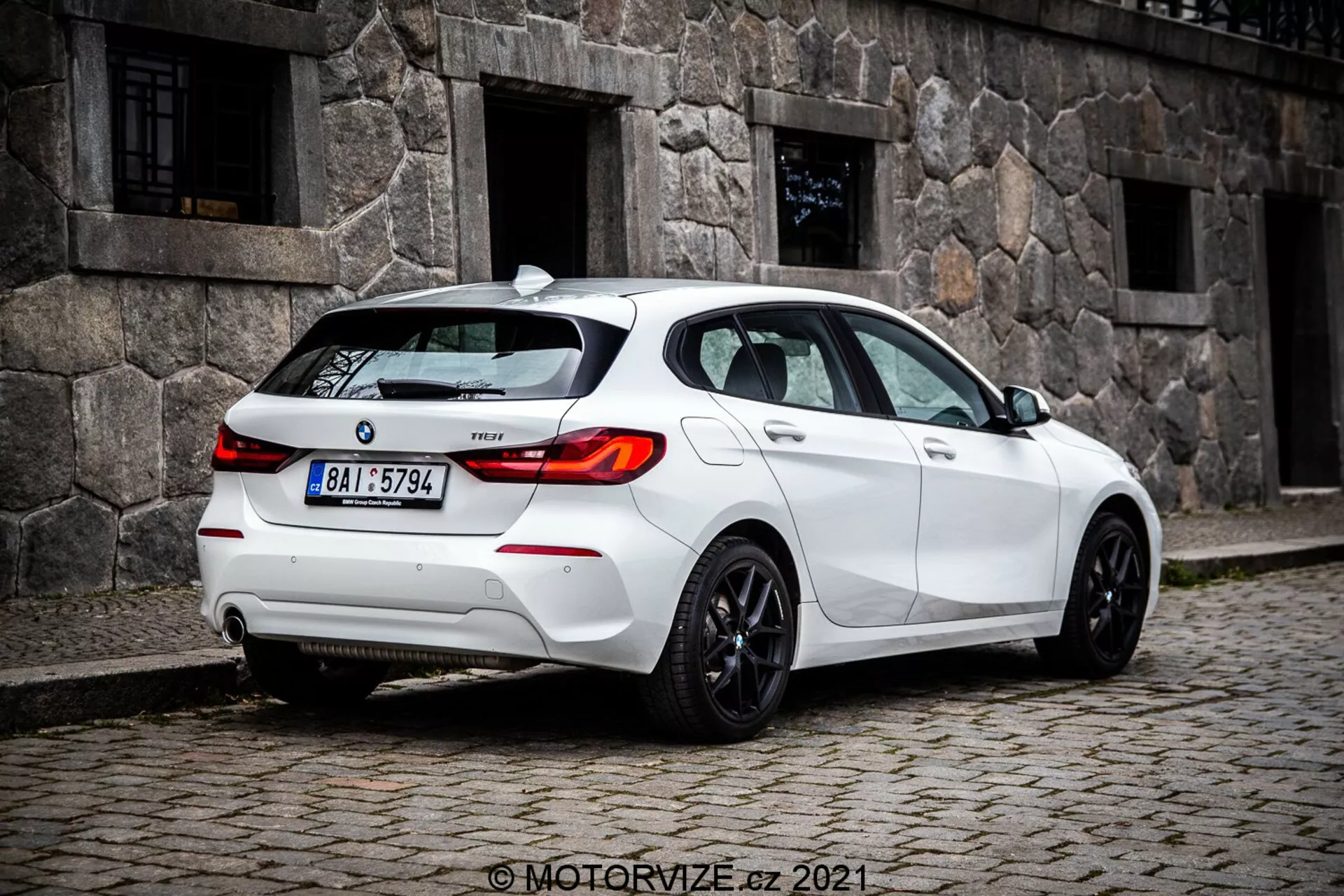 Srednje oddaljen, na nivoju tal posnetek belega hatchbacka BMW 1er-serije 116i 2019, zajet iz tričetrtinskega zadnjega kota, ki izrazito prikazuje zadnje luči z LED obliko, zadnje vetrobransko steklo in športna platišča. Vidne značilnosti vključujejo senzorje na zadnjem odbijaču, verjetno za pomoč pri parkiranju, majhno anteno na strehi, verjetno za GPS ali satelitski radio, oznako modela na prtljažniku, dvojni izpušni sistem, ki nakazuje zmogljivost, in zatemnjena okna za zasebnost ali udobje.