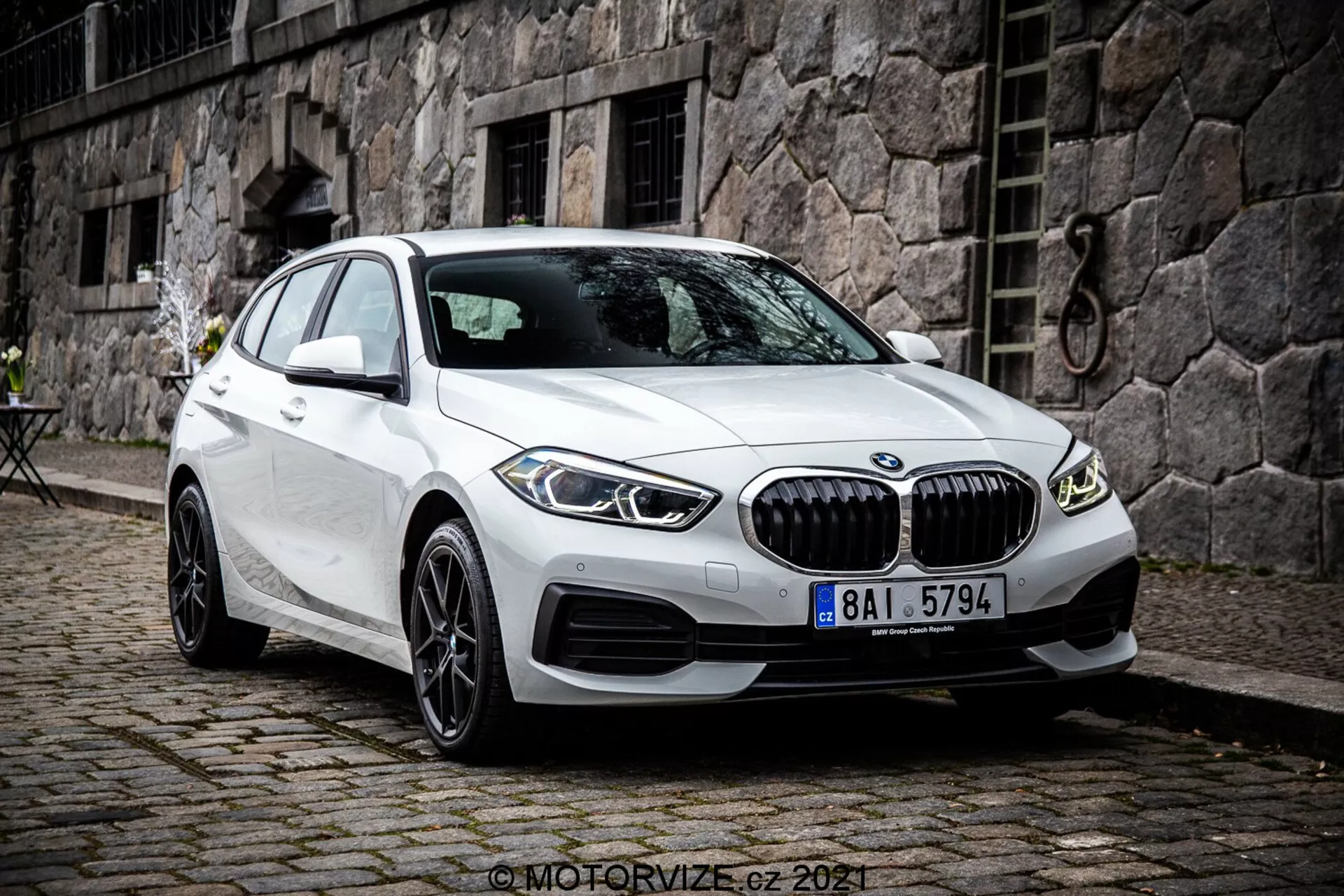 Slika prikazuje bel avtomobil BMW serije 1 iz leta 2019, konkretno BMW 116i. Fotografiran je iz sprednjega trikotnega kota, ki prikazuje prednji del avtomobila, stran potnika in podrobno zasnovo koles. Fotografija je posneta iz rahlo dvignjenega zornega kota, kar nakazuje, da je fotograf stal ali uporabljal stativ. Avtomobil je parkiran s sprednjimi kolesi obrnjenimi v desno, kar omogoča jasen pogled na platišča in stranski profil pnevmatike. Značilnosti vključujejo izrazito sprednjo masko z navpičnimi letvicami, oblikovane linije na pokrovu motorja in stranskih panelih, tanke LED žaromete in dovode zraka na odbijaču, ki lahko vsebujejo parkirne senzorje. Stranska ogledala z vgrajenimi smerniki in brezokvirna okna poudarjajo premium estetiko. Večkraki aluminijasti platišči nakazujejo višjo raven opreme ali opcijsko nadgradnjo. Strmo nagnjeno zadnje okno poudarja profil kombilimuzine. Vozilo je parkirano na tlakovanem pločniku v urbanem ali zgodovinskem okolju s kamnito stavbo in majhno bistro mizo v ozadju. Slika je ostra in podrobna, posneta pri naravni oblačni dnevni svetlobi.