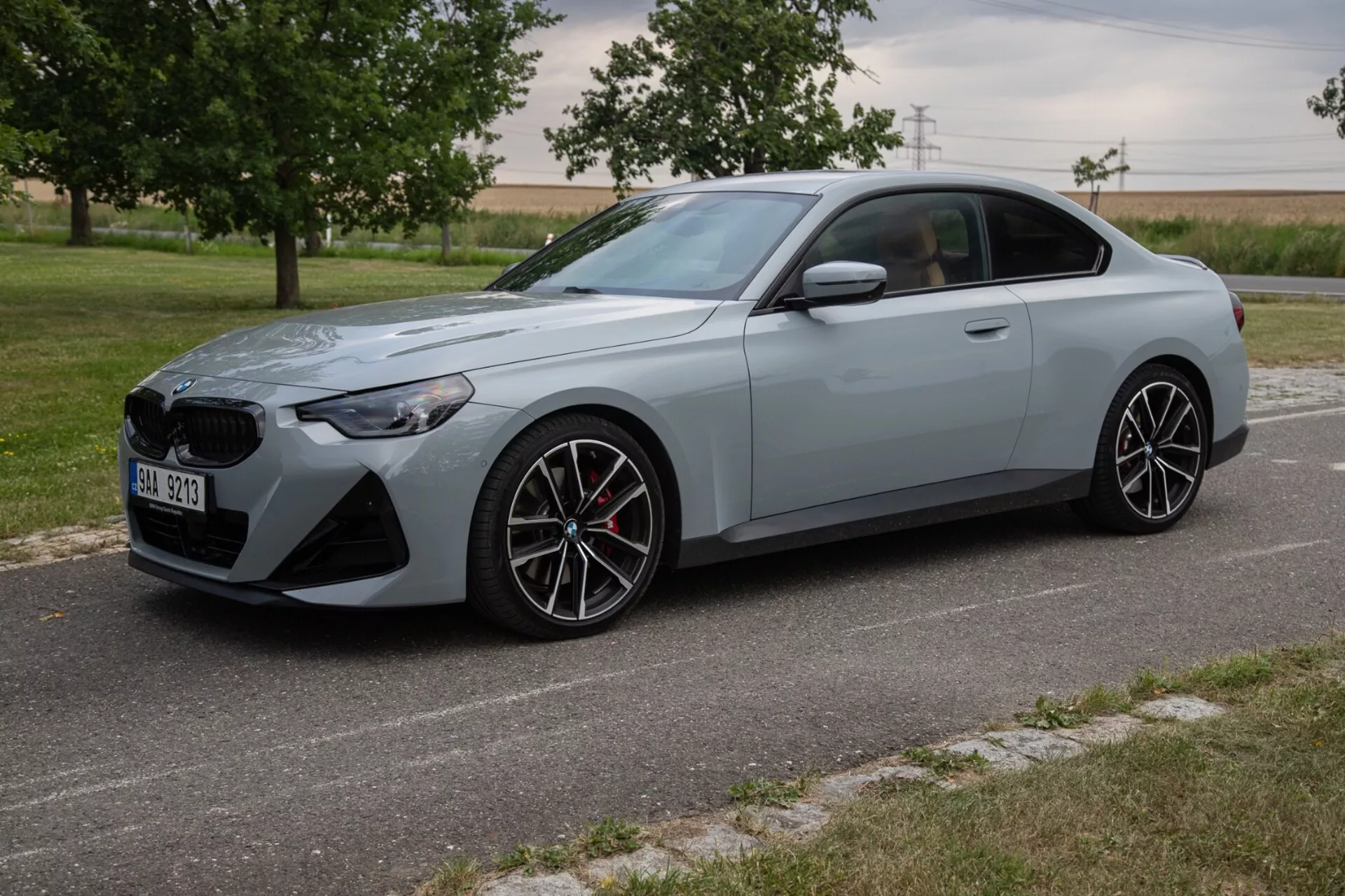 Slika prikazuje BMW 2er-reihe 2022 (2-serija kupe) iz tridimenzionalnega sprednjega kota, ki prikazuje njegovo elegantno in športno zunanjo obliko, z veliko sprednjo masko in kotnimi LED žarometi. Športni kupe, pobarvan v mat svetlo sivo barvo, je opremljen z večkrakimi aluminijastimi platišči, zmogljivim zavorni sistem z rdečimi čeljustmi in izklesanimi telesnimi linijami za aerodinamičen profil. Avto je parkiran na tlakovanem cestišču z naravno pokrajino in polji v ozadju, v dnevnih svetlobnih pogojih.