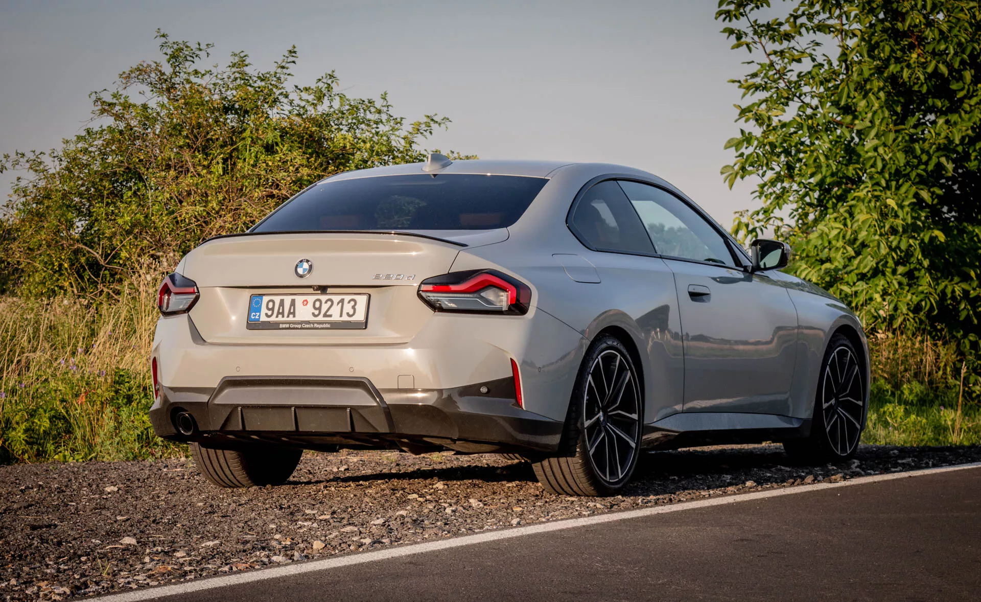 Zadnji pogled na športni avto BMW 2er-reihe 2022 (2-serije coupe) zajet iz nizke perspektive. Avto, parkiran ob cesti v podeželskem okolju z naravno vegetacijo, je viden v mehki svetlobi. Značilne značke označujejo znamko in model, prikazujoc zadnji del, vključno s pokrovom prtljažnika, zadnjim odbijačem in zavitimi zadnjimi lučmi. Dvojne izpušne cevi nakazujejo močan motor. Majhen integriran zadnji spojler in velika aluminijasta platišča s tankimi kraki nakazujeta aerodinamični stil in zmogljivost. Kovinska barva avtomobila in gladko poševna strešna linija povečata njegov premium videz. Vidne možne moderne funkcije vključujejo zadnje LED luči, parkirne senzorje in vzvratno kamero.