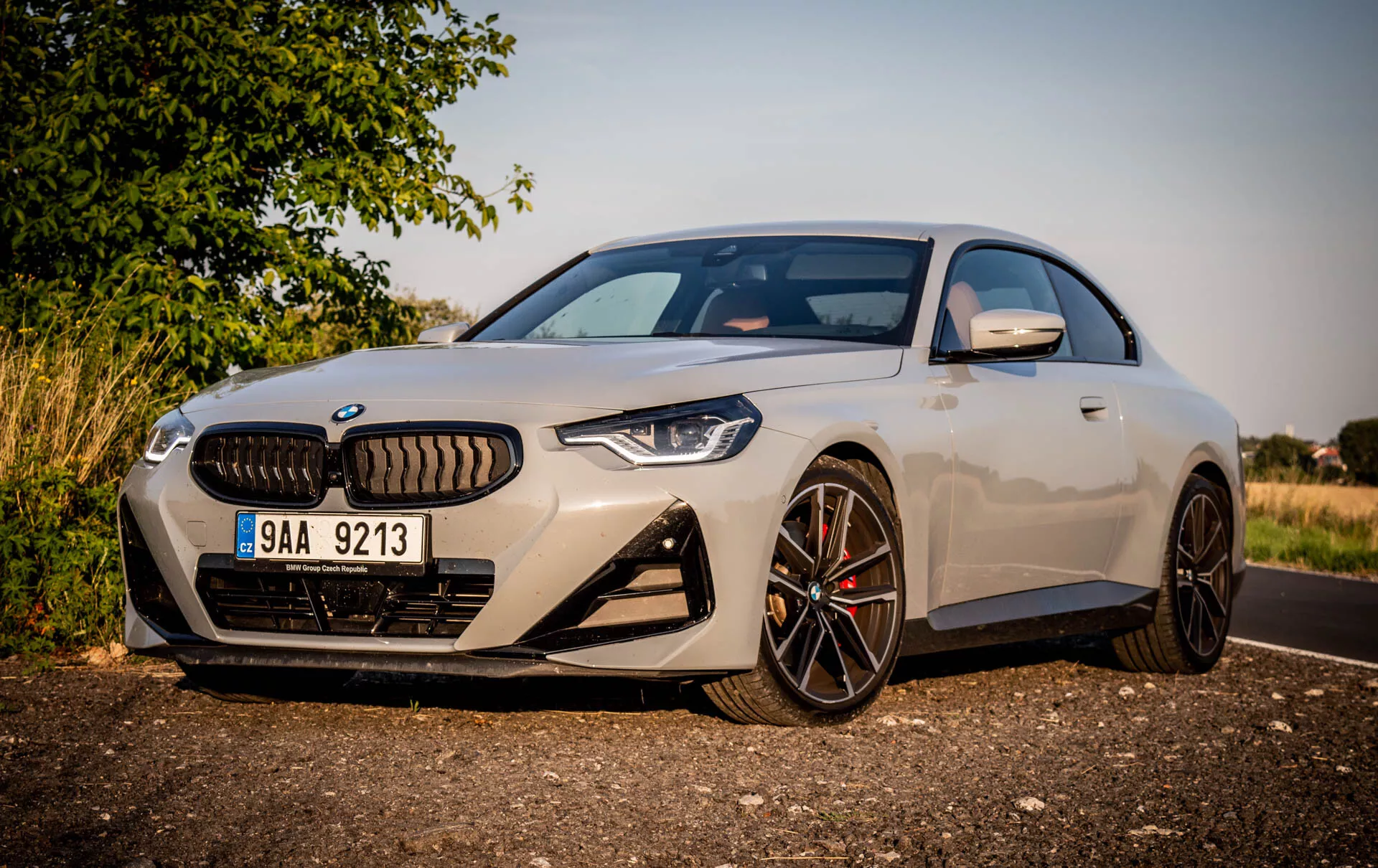 Slika modernega športnega kupeja BMW serije 2 iz leta 2022. Avto je parkiran ob strani ceste podnevi, gledano pod poševnim kotom iz leve sprednje strani, kar poudarja voznikovo stran. Značilnosti vključujejo eleganten aerodinamičen dizajn, velika večkraka lita kolesa s temnim zaključkom, kontrastne zavore, značilno rešetko obdano z ozkimi agresivnimi LED žarometi, sprednjo kamero nad registrsko tablico, velike odprtine za dovod zraka v sprednjem odbijaču, svetlo kovinsko barvo, stranska ogledala z vgrajenimi smerniki ter delni pogled na panoramsko streho. Avtomobil je postavljen blizu kamere, z ruralnim ali primestnim ozadjem, toplim osvetljenjem in sencami, ki nakazujejo nizko sonce bodisi zgodaj zjutraj ali pozno popoldne.
