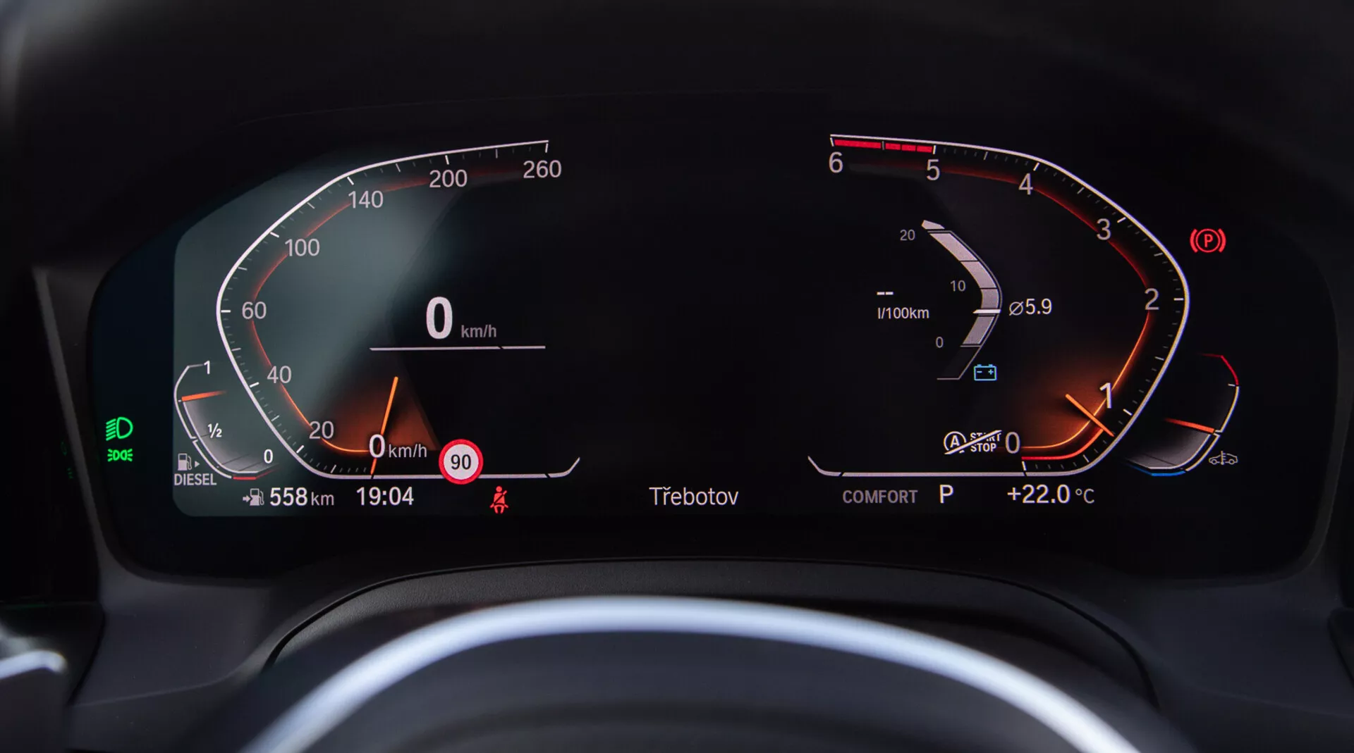Bližnji pogled na digitalno armaturno ploščo BMW 2 Series Coupe 2022, kot jo vidi voznik. Polkrožni digitalni merilnik hitrosti na levi je od 0 do 260 km/h, kar kaže, da vozilo miruje. Na desni strani merilnik vrtljajev prikazuje motor na prostem teku tik nad 0 RPM. V merilnik vrtljajev je integriran skoraj poln digitalni merilnik goriva in merilnik temperature motorja, ki kaže normalen razpon. Osrednji digitalni zaslon prikazuje število prevoženih kilometrov 558 km, hitrost 0 km/h, opozorilo o hitrosti pri 90 km/h, trenutni čas 19:04, indikator prestavne ročice 'P' za parkirni položaj in zunanjo temperaturo +22,0°C. Vidna je tudi zelena ikona asistenčne tehnologije za ohranjanje voznega pasu, prikaz načina 'COMFORT', indikator dizelskega goriva in opomnik za varnostni pas.