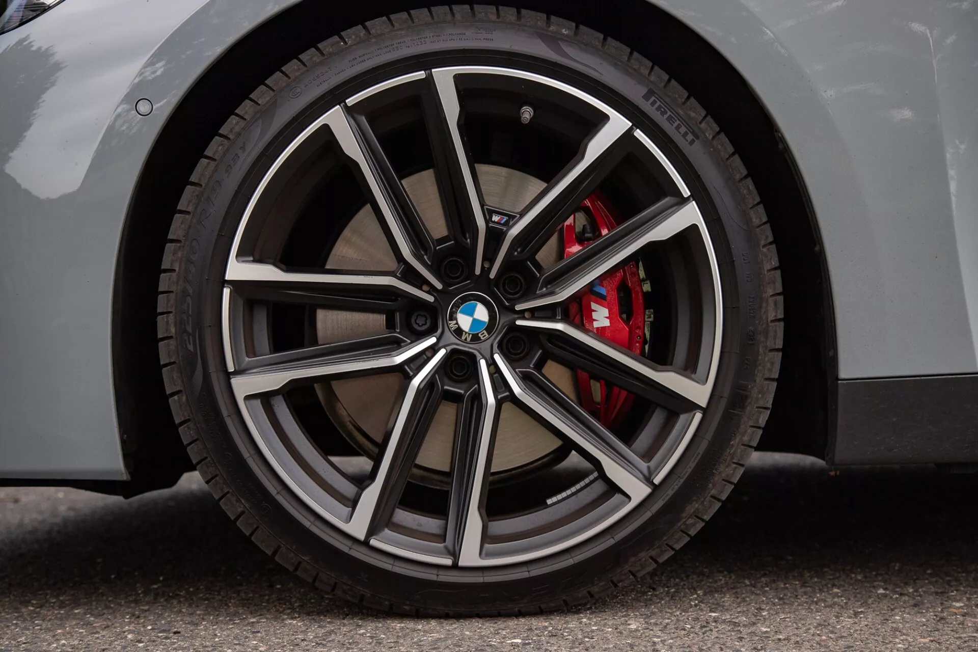 Bližnji posnetek kolesa BMW serije 2 Coupe z večkrako platišče s strojno poliranimi in temnejšimi kovinskimi zaključki, logotipom BMW na pokrovu, nizkoprofilno pnevmatiko in rdečo visoko zmogljivo zavorno čeljustjo. Svetla karoserija na asfaltni površini poudarja luksuzne in športne vidike.