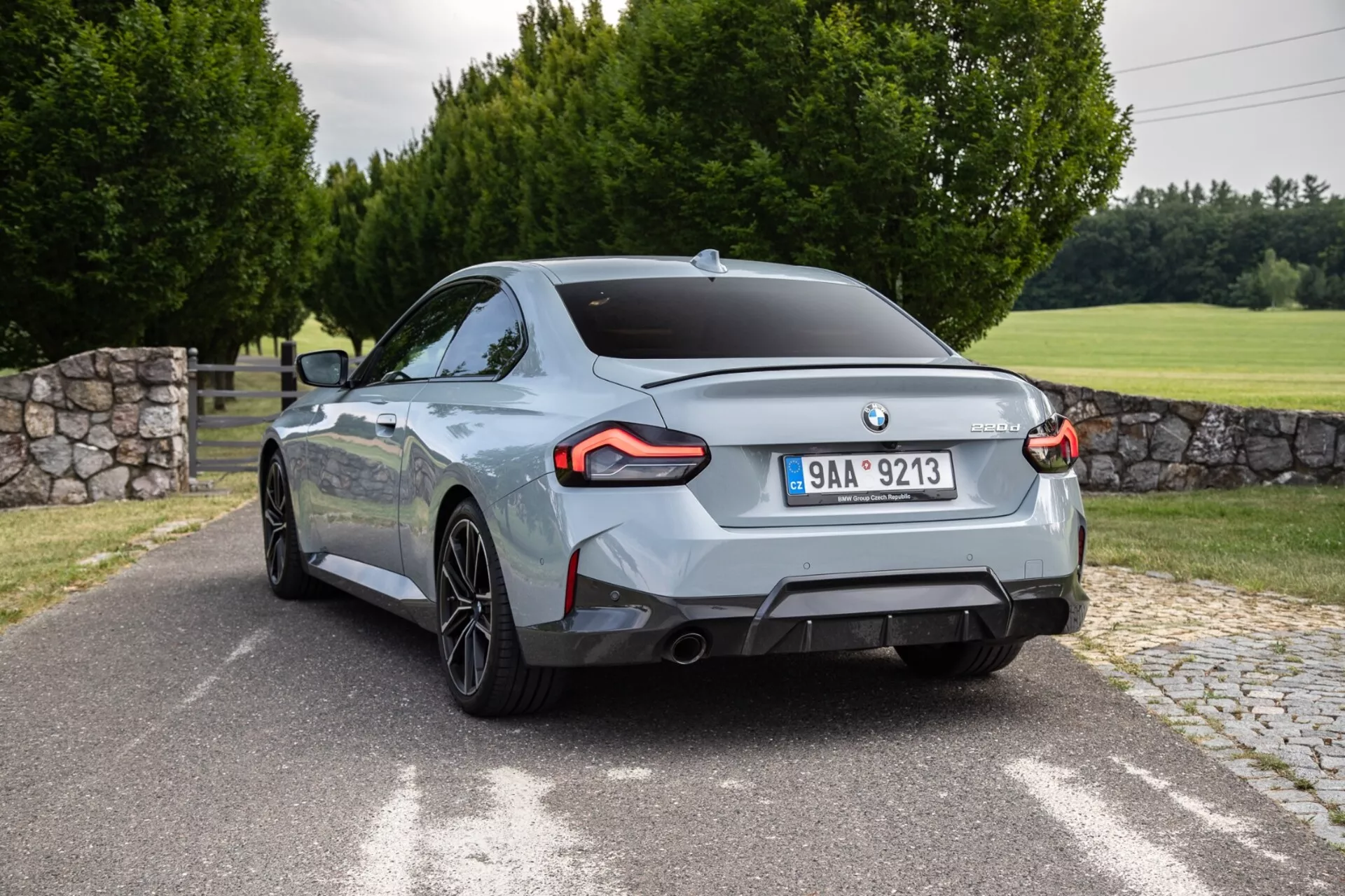 Slika BMW 2er-Reihe 2022 2-Series Coupe, zajeta z zadnjega tričetrtinskega kota. Avto, postavljen skoraj diagonalno, prikazuje značilne zadnje luči LED, dvojne izpušne odprtine, večkratne aluminijaste platišča in sijoč kovinski zaključek barve. Zadnji dizajn vključuje emblem proizvajalca, nameščen na sredini, in značko modela/motorne različice na pokrovu prtljažnika. Zunanja nastavitev vključuje tlakovana cesta ali dovozno pot, travnato površino, drevesa in delno vidno kamnito steno.