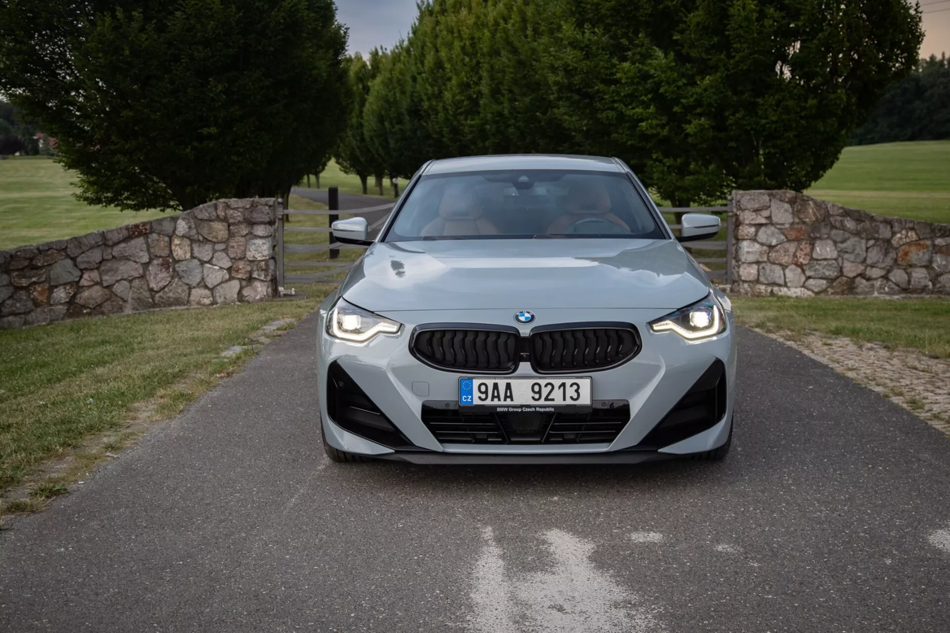 Sprednji pogled na BMW serije 2 2022 (2-series-coupe) v urejenem zunanjem prostoru s značilno rešetko, elegantnimi LED lučmi in aerodinamično oblikovano pokrovom motorja, ki poudarja sodoben dizajn in tehnološke značilnosti, vključno z morebitnimi senzorji za napredne sisteme za pomoč vozniku.