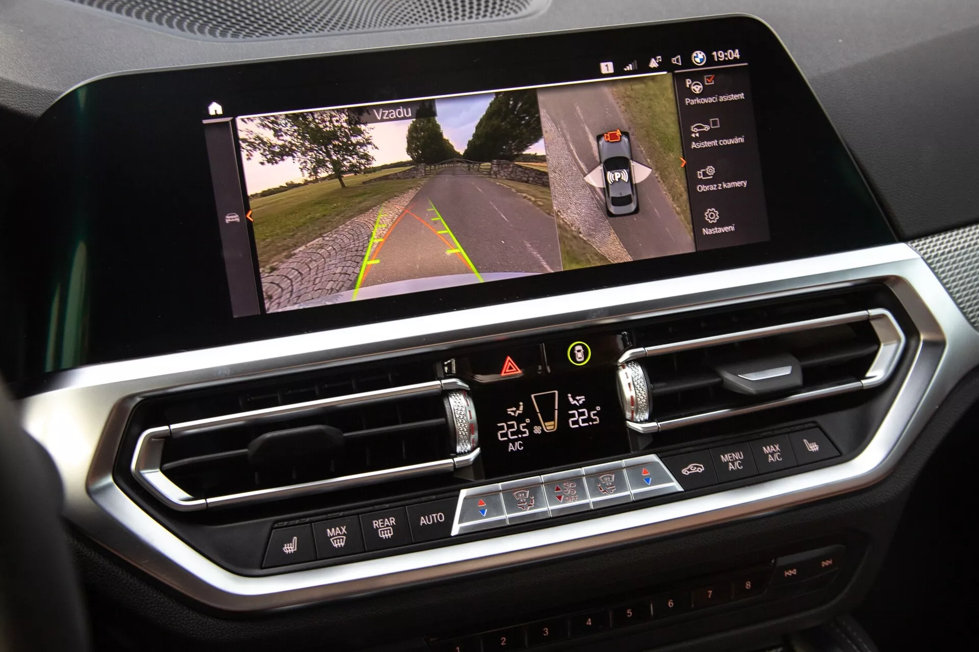 Bližnji posnetek notranjosti BMW 2er-Reihe 2022, ki prikazuje zaslon informacijsko-razvedrilnega sistema in sistem za nadzor klime. Velik, vodoravno usmerjen zaslon na dotik prikazuje pogled na cesto, kar kaže na aktivacijo kamere, medtem ko digitalni vmesnik za nadzor klime spodaj vključuje nastavitve dvojne temperature, hitrost ventilatorja in fizične gumbe. Moderni dizajn s kromiranimi zaključki nakazuje vrhunske materiale.