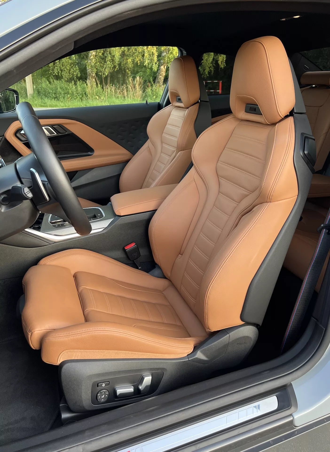 Notranji pogled na BMW serije 2 Coupe 2022 iz voznikove strani vrat, poudarja sprednja sedeža oblazinjena z usnjem v barvi rjavo-karamela s prešitimi podrobnostmi ter ergonomsko podporo. Električne nastavitve sedežev, črn volan s kontrolami ter delni pogled na sredinsko konzolo z menjalnikom, ki nakazuje luksuzne in zmogljivostne značilnosti.