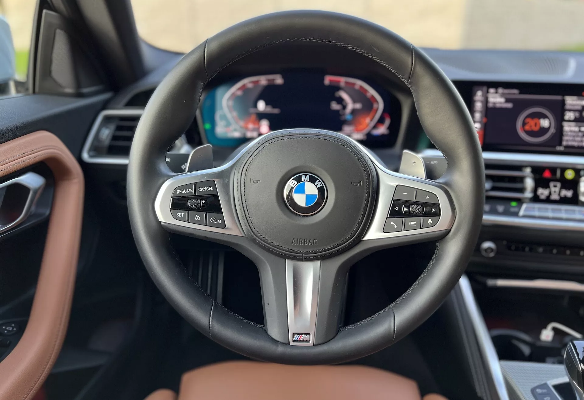 Notranji posnetek BMW 2er-Reihe 2022, osredotočen na volan in armaturno ploščo iz perspektive voznikovega sedeža. Vsebuje trikraki volan BMW z večfunkcijskimi gumbi za tempomat, upravljanje zvoka in telefonijo, skupaj z obvolanskimi ročicami in oznako zmogljivosti 'M'. Za volanom je digitalna instrumentna plošča, ki prikazuje tahometer, hitrostomer in večfunkcijske informacije. Sistem informacijske zabave je viden na sredini armaturne plošče, notranja obloga pa vključuje usnje in les, kar kaže na vrhunski model.