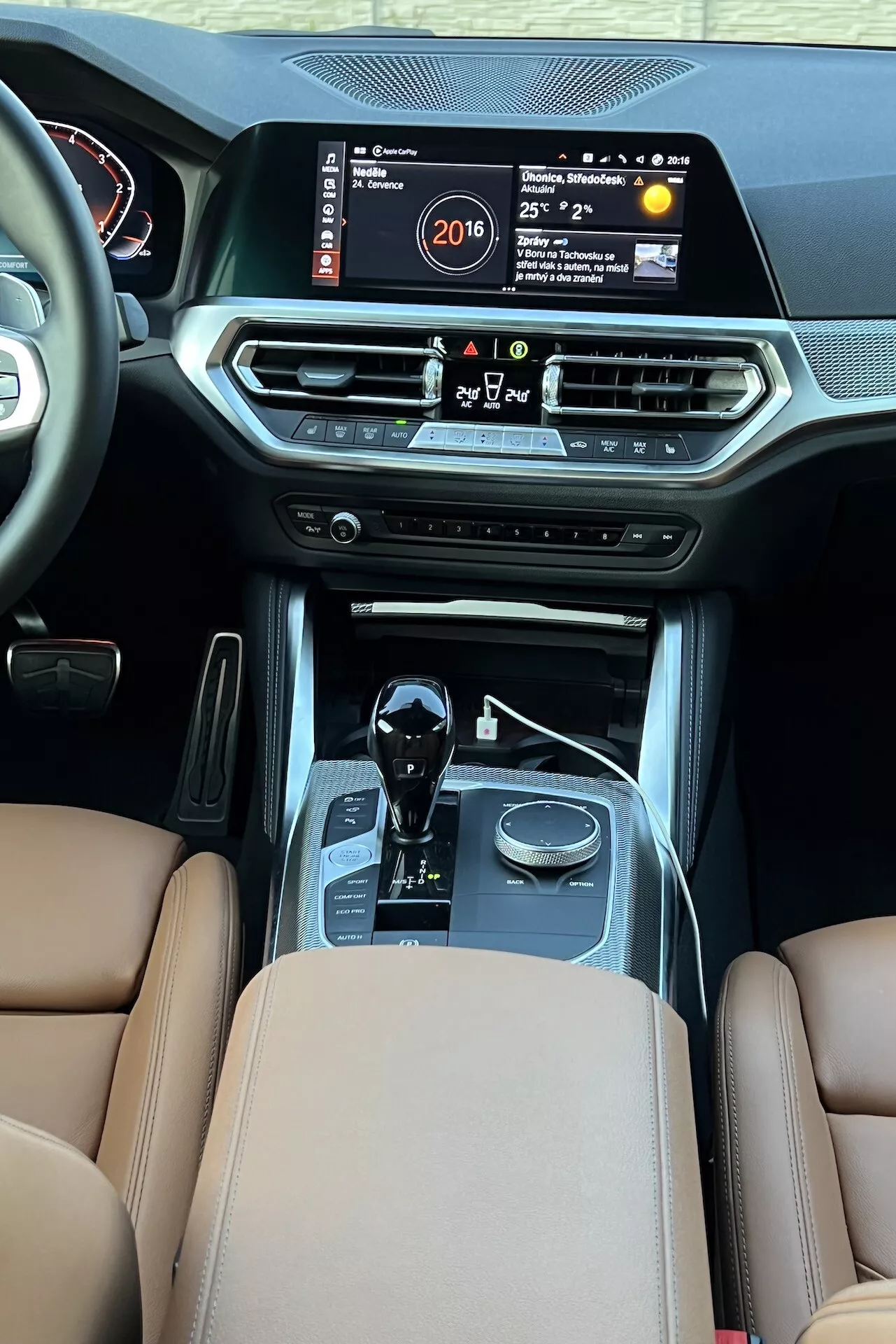 Notranji pogled na BMW serije 2 2022 iz zadnjega potniškega prostora, ki poudarja osrednjo konzolo z velikim informacijsko-zabavnim zaslonom, ki prikazuje čas, temperaturo in multimedijski sistem. Lastnosti vključujejo prezračevalne reže za klimatsko napravo, gumbe za nadzor klime, zagonski gumb, prestavno ročico samodejnega menjalnika s kromiranimi poudarki, izbirnike načinov vožnje, rjave usnjene sedeže s premijskim oblazinjenjem in šivi, kar nakazuje na luksuz in napredno avtomobilsko tehnologijo.