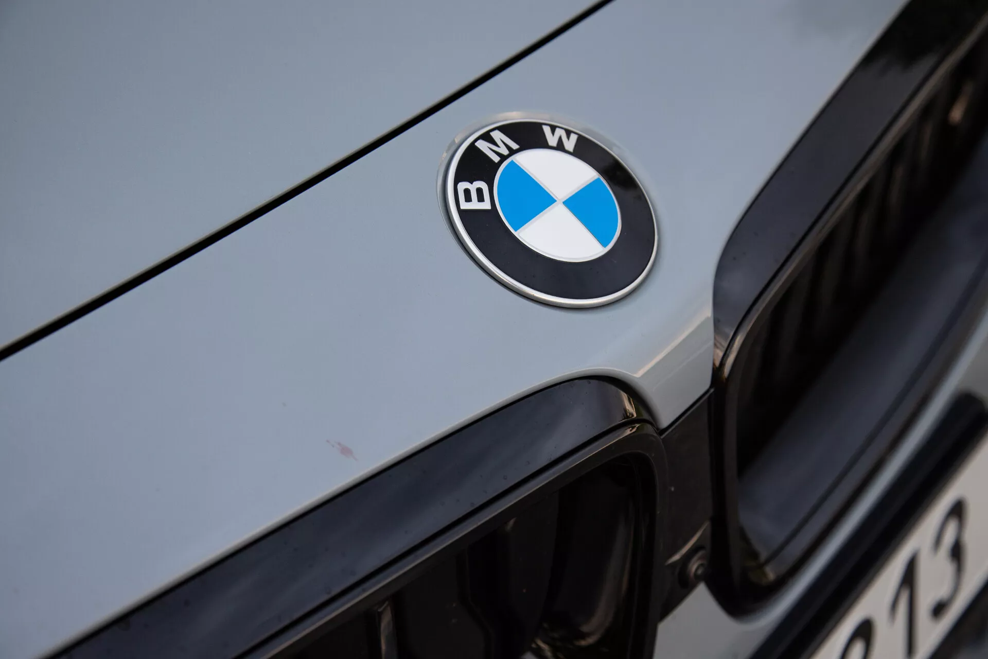 Bližnji posnetek sprednjega dela BMW 2-Series Coupe 2022, z jasno centriranim črnim, modrim in belim BMW-jevimi znaki na pokrovu motorja. Slika prikazuje zgornji rob ikonične ledvične mreže v ozadju, s kovinsko srebrno ali svetlo sivo avtomobilsko zunanjostjo, ki odbija ambientalno svetlobo. Ostri fokus na znak poudarja luksuzno blagovno znamko in dobro vzdrževano površino vozila.