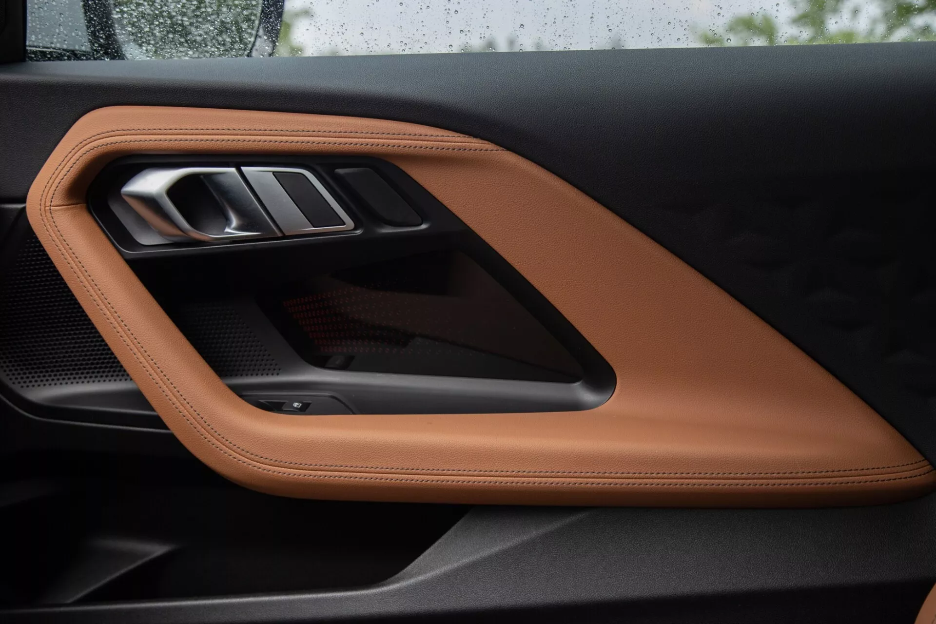 Od blizu posnetek notranje plošče vrat BMW 2 Series Coupe 2022, ki prikazuje razkošno šivano usnje v barvi svetlo rjave ali konjak, elegantno kovinsko ročico vrat in športni karbonski poudarek, ki poudarjajo visokokakovostne materiale in prefinjene oblikovalske elemente.
