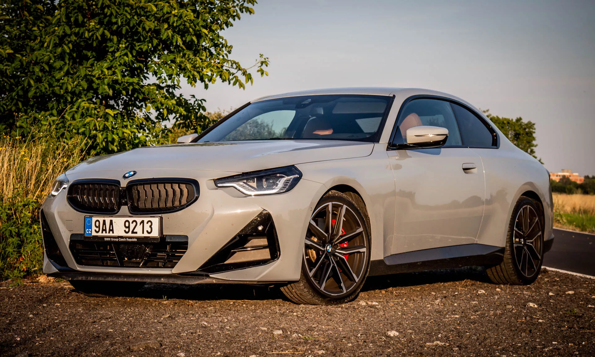 Slika BMW serije 2 coupe 2022. Moderni luksuzni avto je parkiran pod kotom, poudarjen je levi sprednji del med zlato uro. Posnetek se osredotoča na značilne oblikovne elemente, kot so velika ledvična mreža, kotni LED žarometi, športni sprednji odbijač z velikimi zračnimi odprtinami in črna platišča iz več krakov s visokozmogljivimi zavorami. Aerodinamično stransko ogledalo ima vgrajene smernike. Elegantna karoserija in kakovostna barvna obdelava poudarjata kompakten, športen profil avta in nizko strešno linijo.
