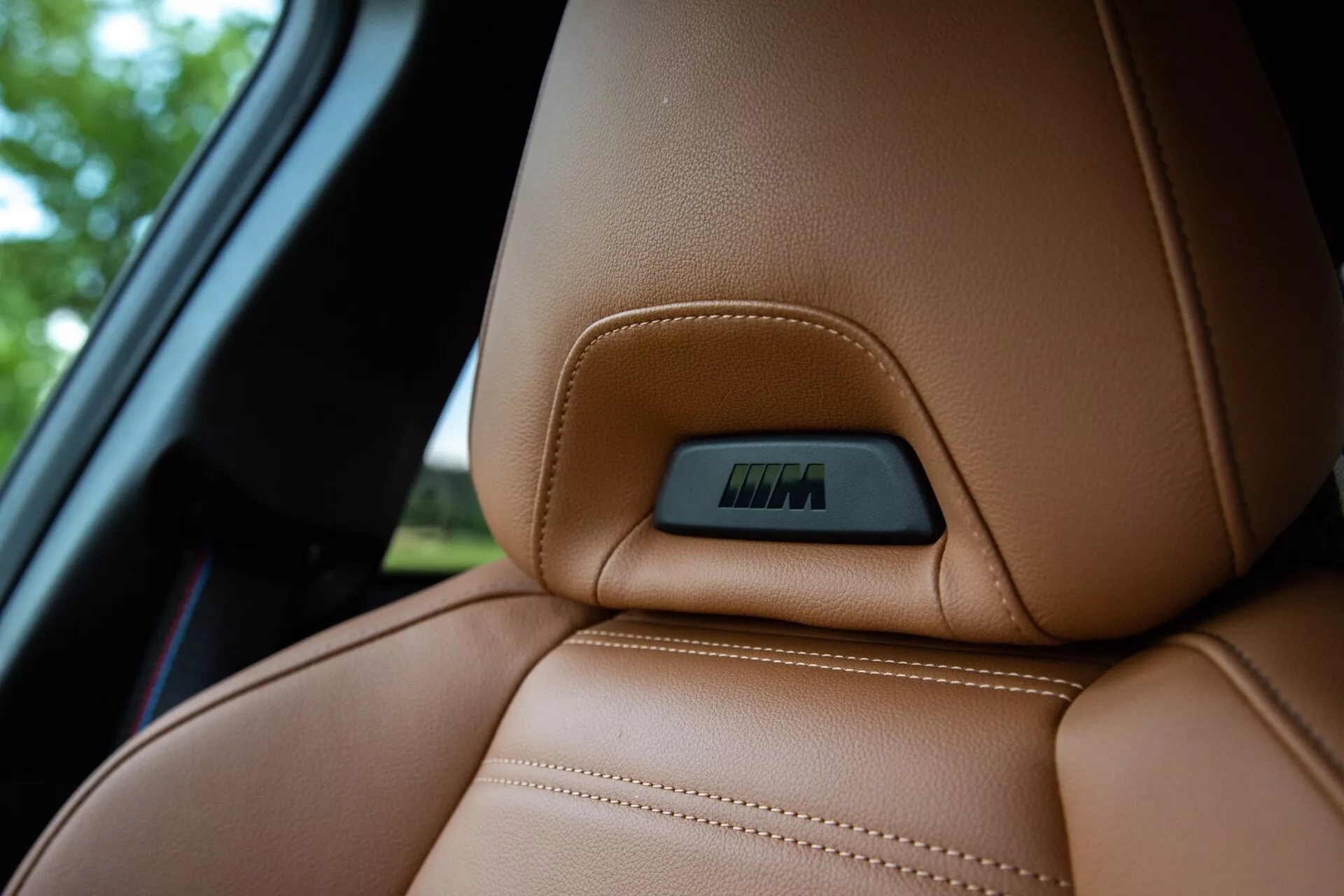 Prikaz od blizu zgornjega dela rjavega usnjenega sedeža v BMW 2 Series Coupe 2022, poudarja natančne vidne šive in ergonomsko zasnovano linijo kontur za udobje in podporo. Na zgornjem delu sedeža je vgrajena značka, ki označuje tehnološke funkcije, kot so elektronske nastavitve ali ogrevalne funkcije. Posnetek je narejen iz nekoliko nižje perspektive, poudarja razkošno teksturo kože in zamegli ozadje, ki vključuje del avtomobilskega okna.