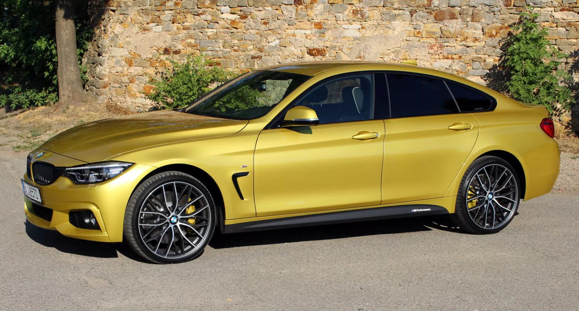 Metalik zlato kupé BMW serije 4 iz leta 2013 parkirano vzporedno s starim kamnitim zidom, prikazuje svoj eleganten, aerodinamičen stranski profil, zlitinska kolesa in tehnološke izboljšave z rahlo dvignjenega kota
