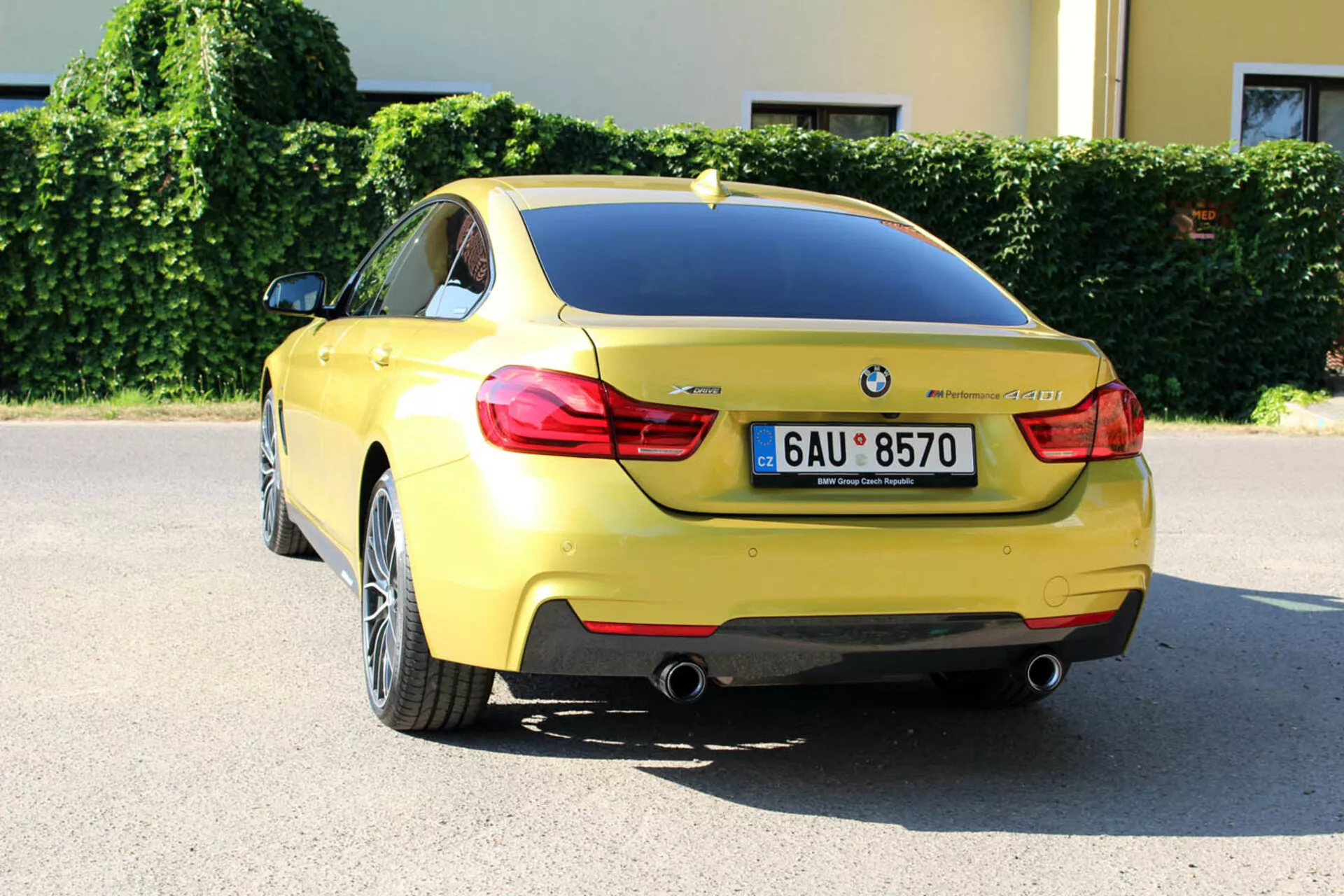 Rumeni BMW serije 4 Coupe letnik 2013 iz zadnjega kota tri četrtine prikazuje obliko prtljažnika, zadnji odbijač, del zadnjega kolesnega ohišja, značilne zadnje luči, dvojne izpušne cevi, registrsko tablico, značko BMW, oznako modela, parkirne senzorje, anteno v obliki morskega psa in večkraka lita kolesa, parkiran v mestnem okolju na sončen dan.