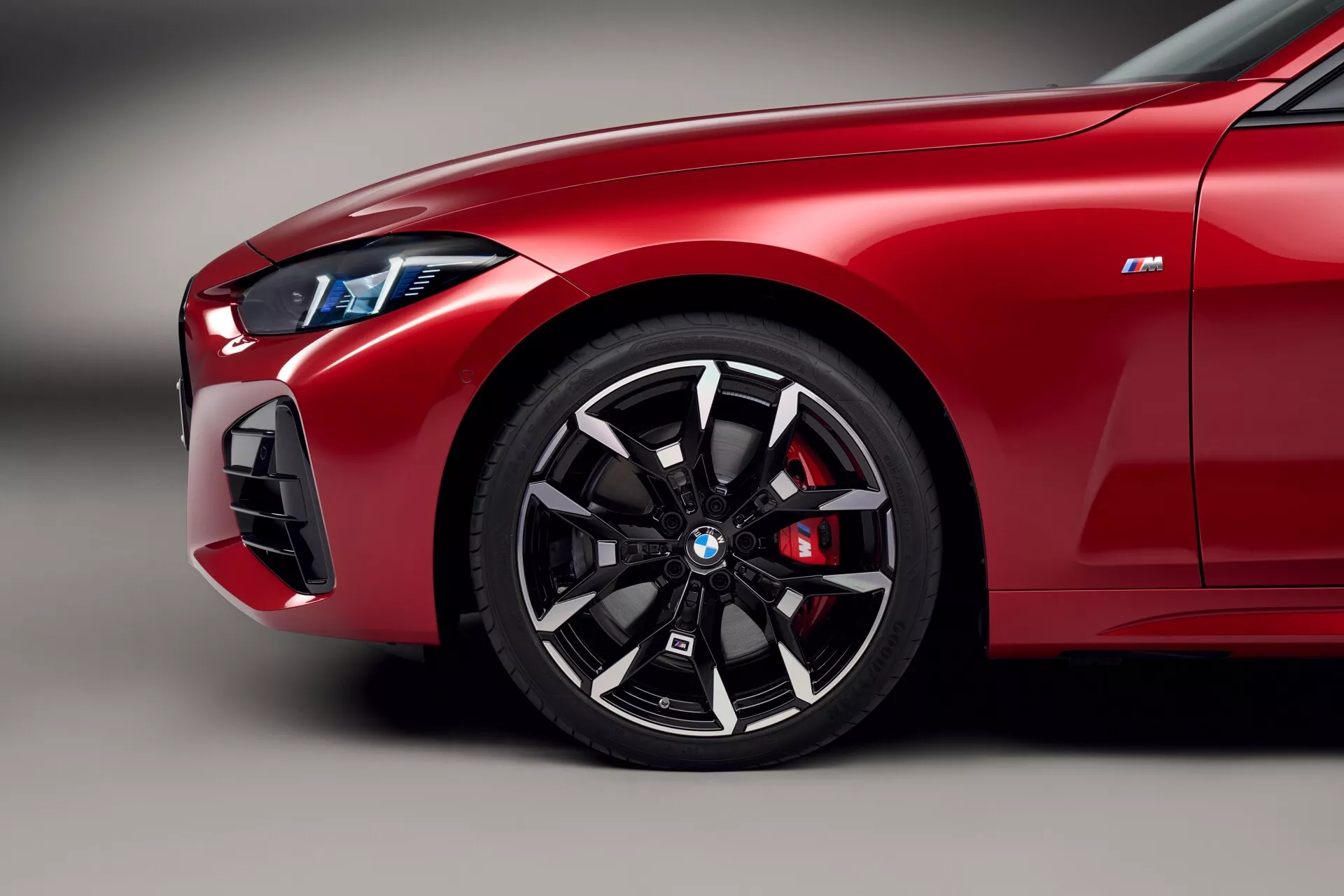 Podrobna slika rdečega sprednjega četrtletja BMW serije 4 (G22-G23-G26-G82-G83 prenova) za leto 2024, ki prikazuje večkrako kolo, visokozmogljivi zavorni čeljust, elegantno LED žaromet in značko 'M', ki nakazuje značilnosti serije BMW M.
