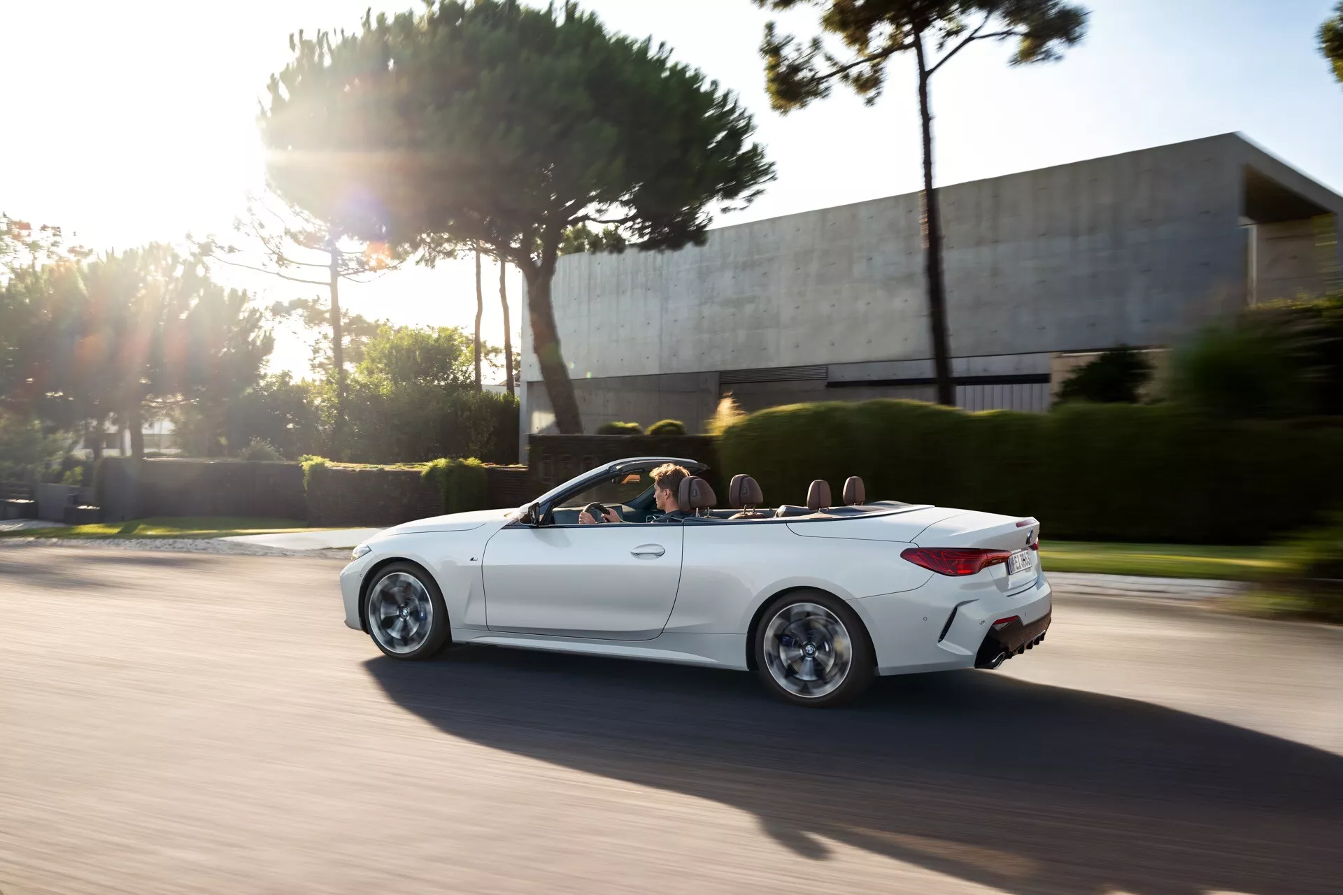 BMW serije 4 Cabrio (G22-G23-G26-G82-G83 Facelift) 2024 v gibanju, ki prikazuje svojo belo elegantno zasnovo z izrazitimi kolesnimi lokarji, stilnimi zlitinimi kolesi in jasnim pogledom v svojo razkošno notranjost preko mehanizma zložljive strehe, med zlato uro s dinamično, športno držo poudarjeno z zameglitvijo hitrosti in sončnim bleščanjem.