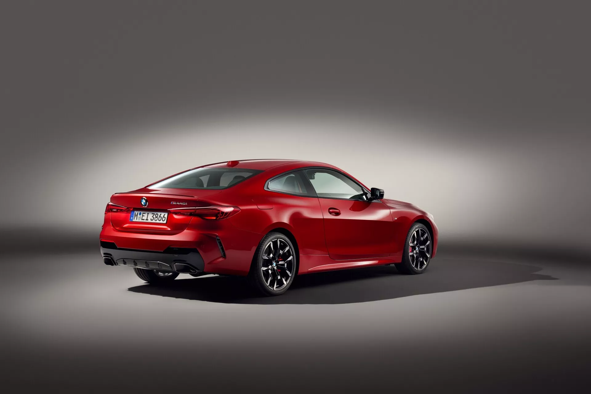 Rdeč športni kupe BMW serije 4 (G22-G23-G26-G82-G83 Facelift) iz leta 2024 v treh četrtinskih zadnjem pogledu proti gradientno sivemu ozadju, ki prikazuje njegov aerodinamični dizajn, mišičasti zadnji kolesni lok, večkraka aluminijasta kolesa, tanke horizontalne zadnje luči, dvojne izpušne konice, vgrajeni zadnji spojler in LED tehnologijo na nizkoprofilnih zmogljivih pnevmatikah, kar odraža poudarek na luksuzu in visoki zmogljivosti.