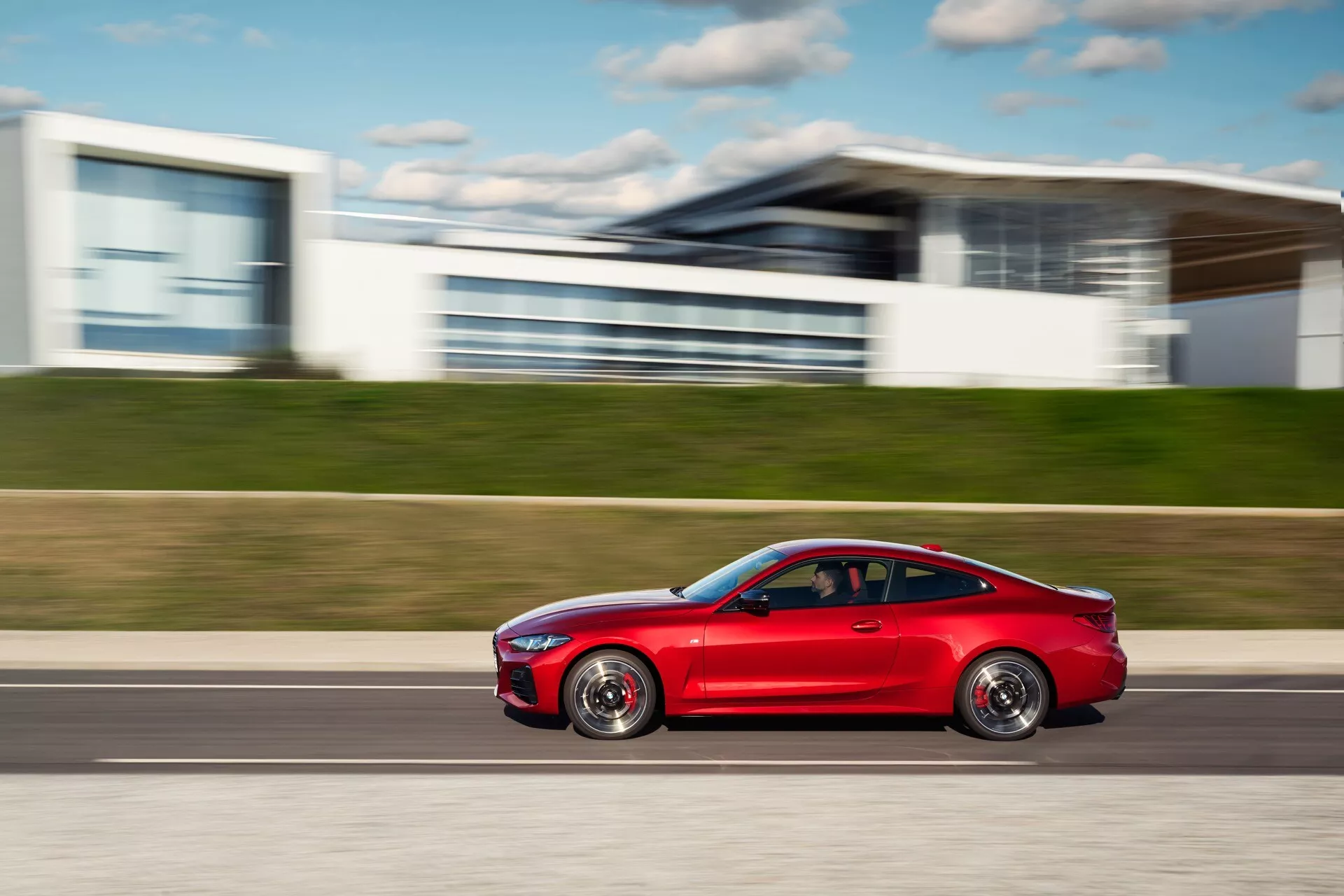 Moderen rdeč kupe BMW serije 4 (G22-G23-G26-G82-G83 Facelift) v gibanju, z aerodinamično zasnovo, športnimi platišči iz zlitine, toniranimi okni in zmogljivimi pnevmatikami, zajet v dinamičnem stranskem posnetku z zamegljenim urbanim ozadjem.