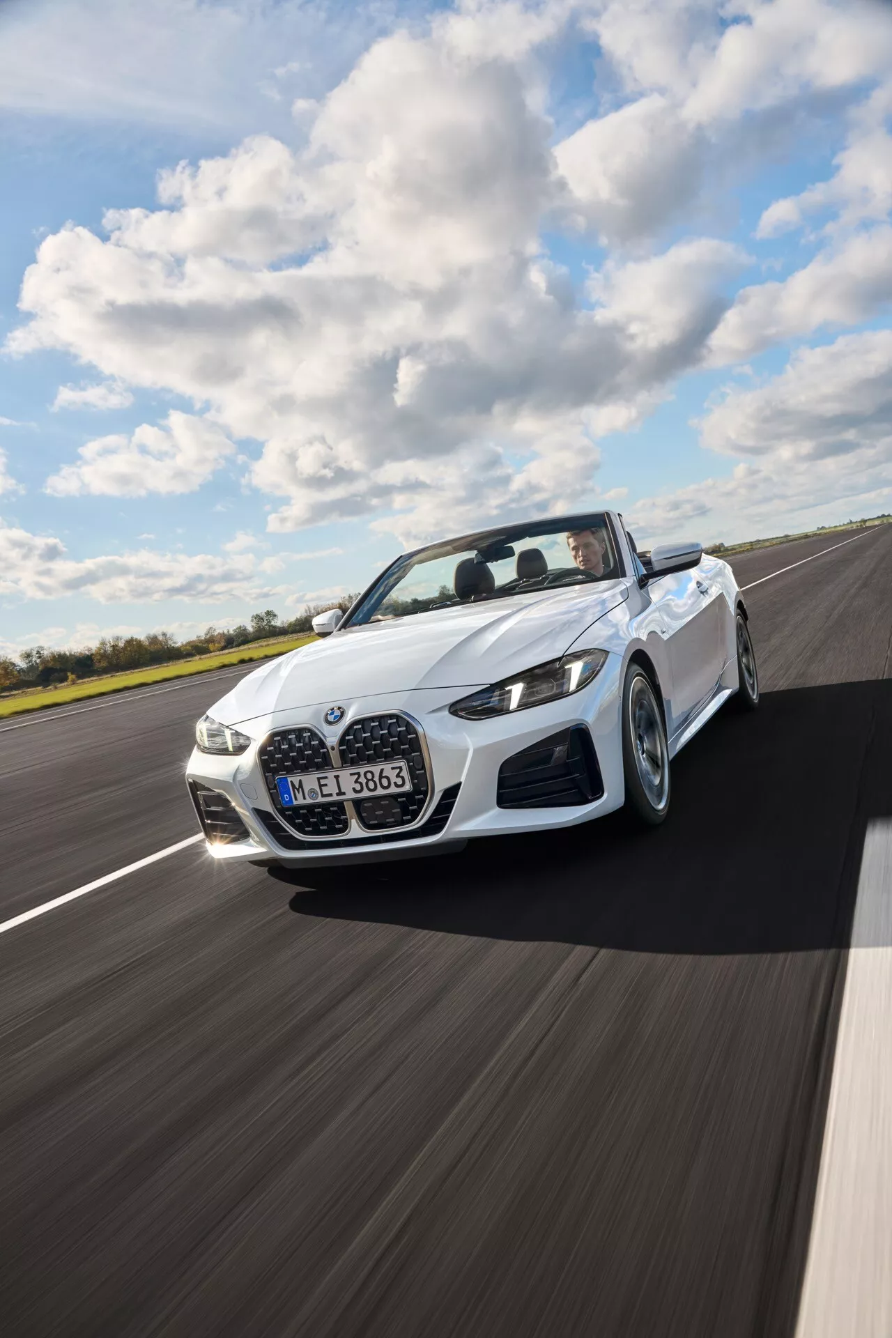 BMW serije 4 kabriolet (G22-G23-G26-G82-G83 Facelift) letnik 2024 v beli barvi, vozeč se po suhi asfaltni cesti pod delno oblačnim nebom, prikazuje dinamičen pogled s treh četrtin