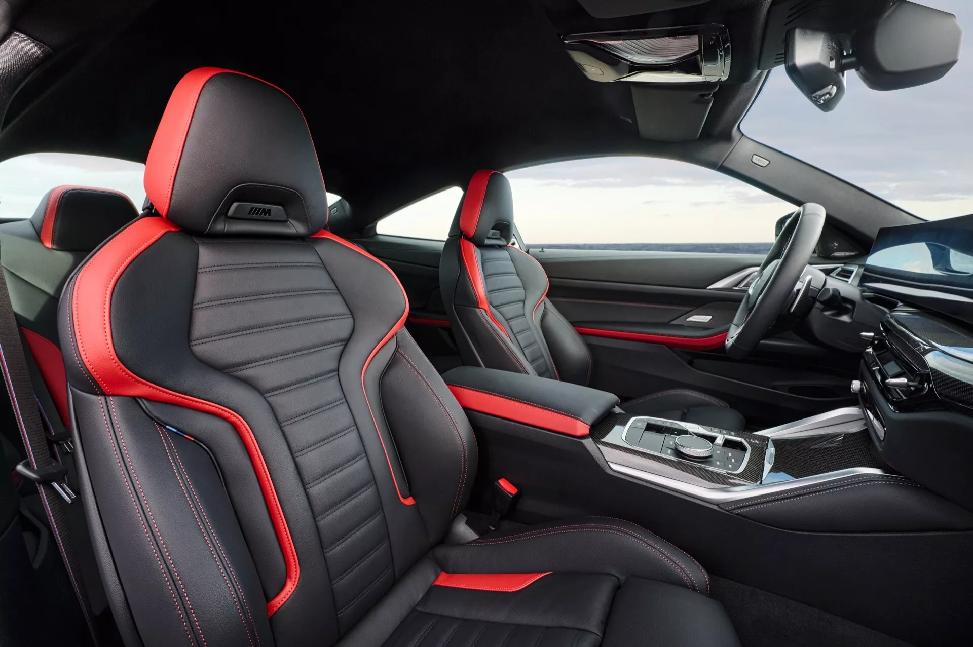 Notranji pogled na BMW serije 4 letnik 2024 (G22-G23-G26-G82-G83 Facelift), ki izpostavlja športno oblikovane sprednje sedeže iz črnega usnja z rdečimi šivi, elegantno osrednjo konzolo z metalnimi poudarki, napreden zaslon armaturne plošče z infotainment sistemom in moderno ergonomsko oblikovanje