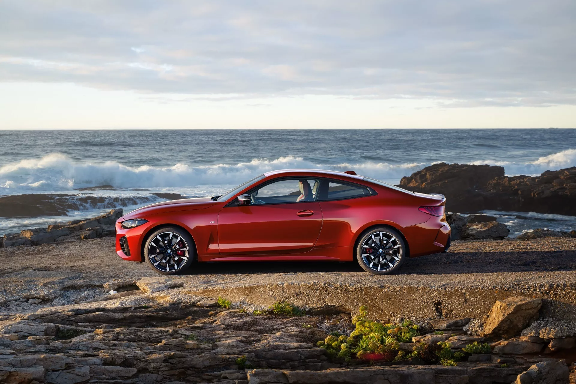 Kupe BMW serije 4 (G22-G23-G26-G82-G83 Facelift) letnik 2024 v svetle metalik rdeči barvi, parkiran ob skalnati obali z valovi in oceanom v ozadju pod delno oblačnim nebom, ki razkriva svoj športni stranski profil z velikimi večkrakimi aluminijastimi kolesi in elegantno aerodinamično zasnovo