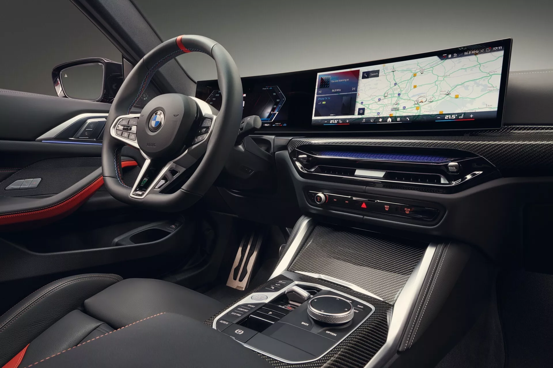 Pogled na notranjost BMW serije 4 letnik 2024 (G22-G23-G26-G82-G83 Facelift), ki prikazuje značko na športnem, multifunkcijskem volanu, digitalno armaturno ploščo z raztegnjenim zaslonom, elegantno sredinsko konzolo z visokim sijajem in sedeže z dvobarvno barvno shemo z rdečimi poudarki.