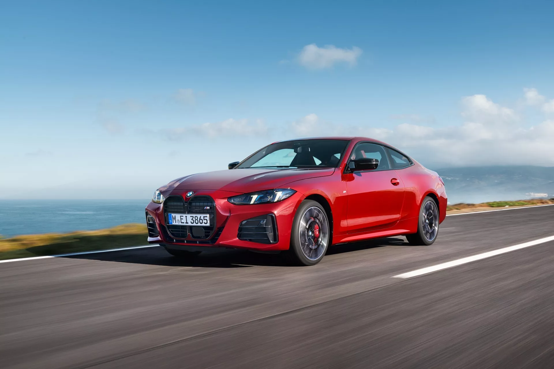 Živo rdeče kupé BMW serije 4 iz leta 2024 (G22-G23-G26-G82-G83-Preoblikovanje) s športno držo na obalni cesti, ki prikazuje aerodinamično oblikovanje, elegantne LED žaromete in značilno rešetko