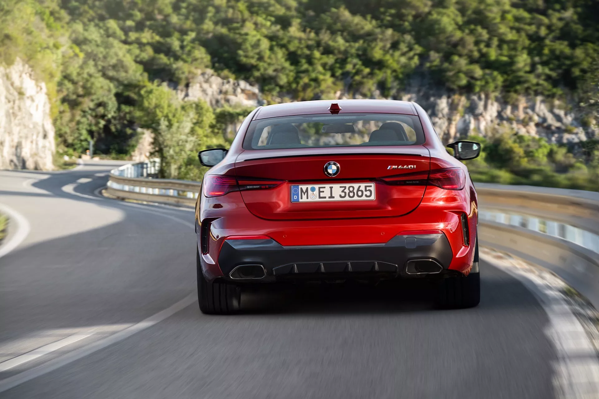 Zadnji pogled na rdeč BMW M8 Competition v gibanju, ki prikazuje BMW emblemo, oznake M8 in serije M, edinstvene zadnje luči, štirikratne izpušne cevi in aerodinamični zadnji difuzor na podeželski cesti z naravnim ozadjem.