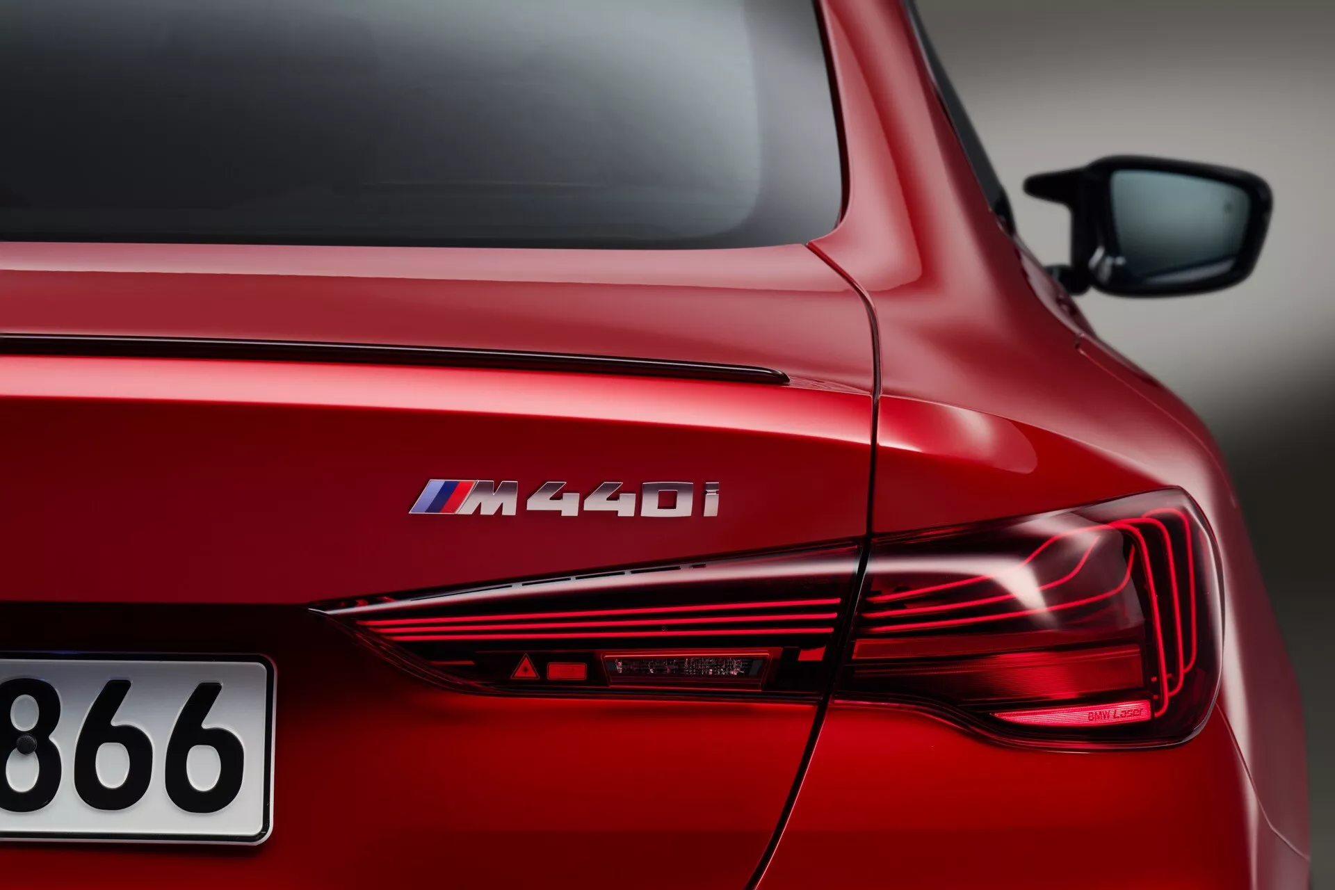 Iz bližje perspektive, poševni pogled na zadnji del rdečega BMW serije 4 2024 (G22-G23-G26-G82-G83-Facelift) M440i, ki prikazuje značilno oznako modela, elegantne LED zadnje luči, kiparsko zunanjost in sijočo kovinsko rdečo končno obdelavo pod umetno osvetlitvijo.