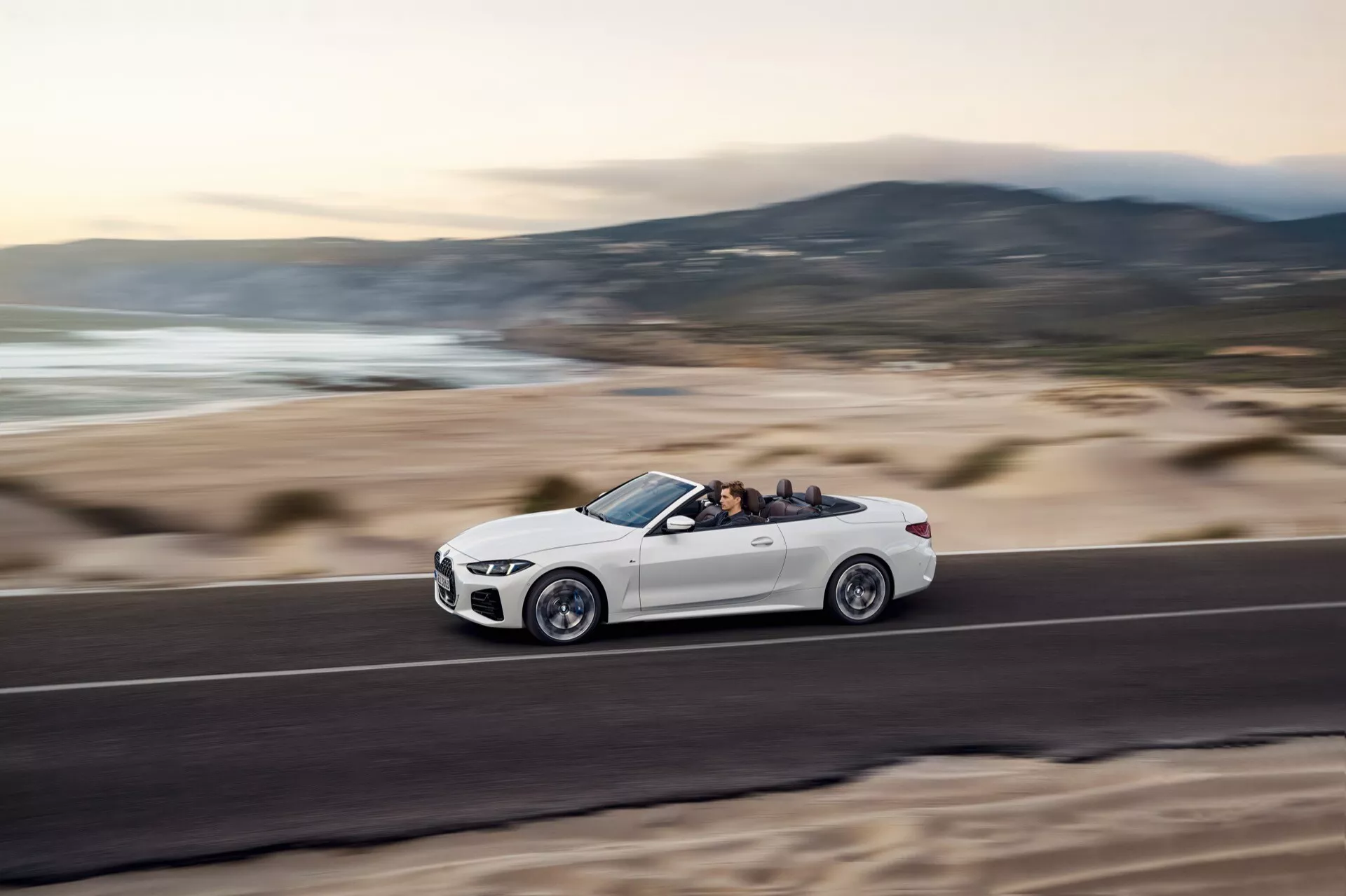 BMW Serije 4 Kabriolet 2024 (G22-G23-G26-G82-G83 Facelift) v gibanju na obalni cesti, prikazuje stranski profil z dinamično držo zaradi posnetka iz nizkega kota, vidni detajli koles in opreme, spuščena streha razkriva notranjost in potnike, proti zamegljenemu ozadju plažne pokrajine