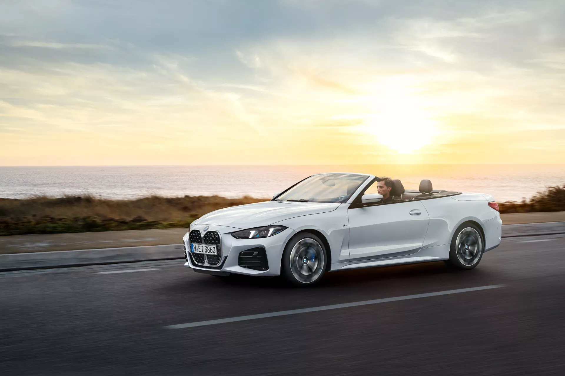 BMW serije 4 kabriolet (G22-G23-G26-G82-G83 Facelift) iz leta 2024 v gibanju na obalni cesti ob sončnem vzhodu ali zahodu, prikazuje svoje aerodinamične karoserije, zlitine koles, LED žaromete, luksuzno notranjost in napredno tehnologijo