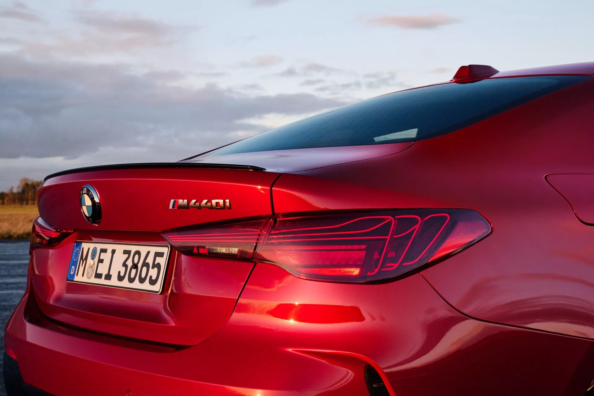 Zadnji del BMW serije 4 2024 (G22-G23-G26-G82-G83 Facelift) pogledan z visokega kota tri četrt, prikazuje desni zadnji kvartal, dizajn LED zadnjih luči, integriran zadnji difuzor, del zadnjega okna in C-stebrička, s poudarkom na elementih dizajna luksuznega športnega sedana, oznakah zmogljivosti in metalik rdeči barvi v zunanji nastavitvi ob sončnem vzhodu ali zahodu.