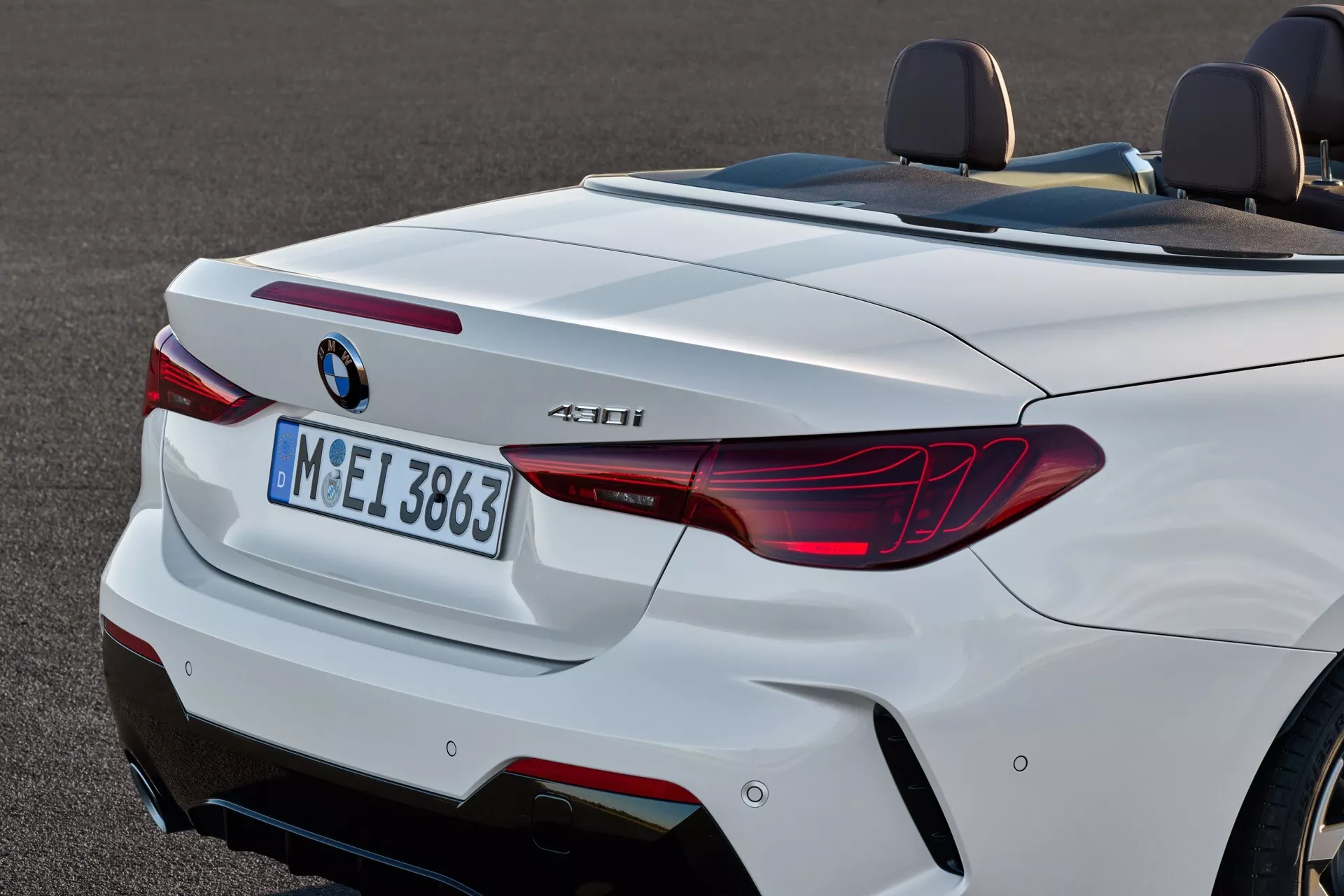 Zadnji pogled na beli kabriolet BMW serije 4 (preoblikovanje 2024) z LED lučmi v obliki L, dvojnimi izpušnimi cevmi in športnim difuzorjem, s poudarkom na aerodinamični zadnji silhueti in tehnoloških značilnostih, kot so senzorji za parkiranje zadaj