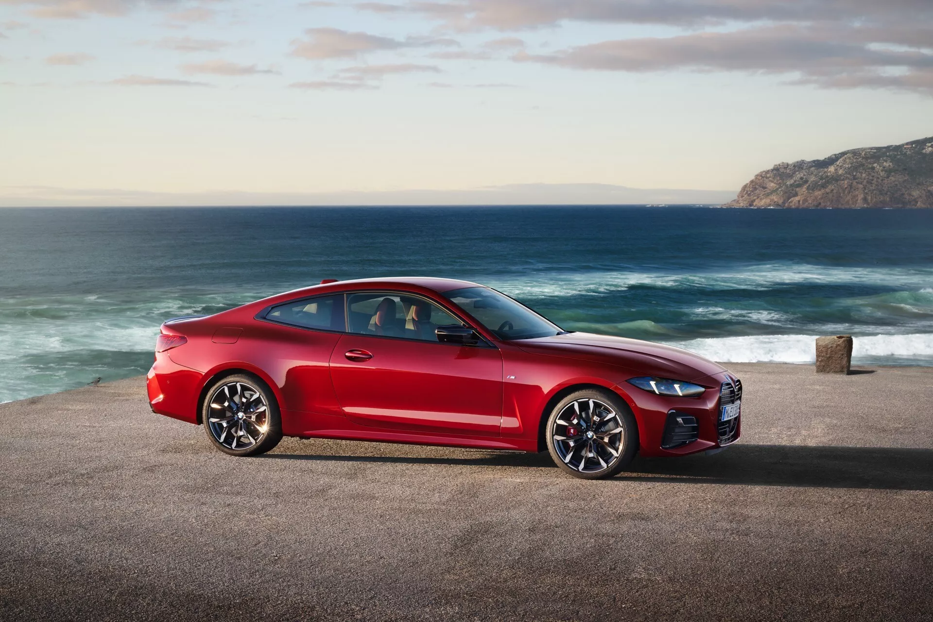Rdeč športni kupe BMW serije 4 (preoblikovanje G22-G23-G26-G82-G83) 2024 parkiran na obalni cesti z oceanom in hribovito obalo v ozadju, ki prikazuje bočni profil vozila, elegantno zasnovo in zmogljivostne funkcije v naravni dnevni svetlobi
